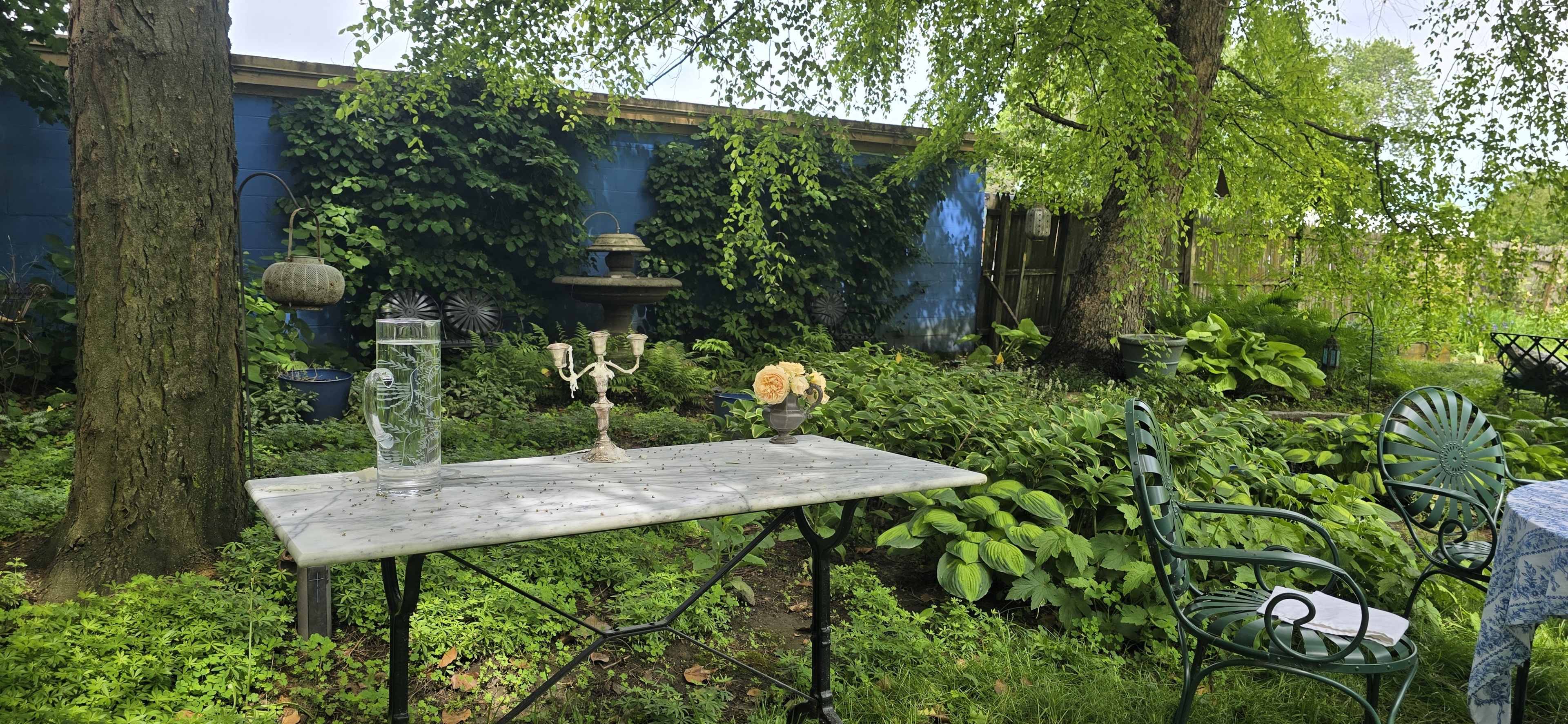 A marble table is set in a lush garden with green foliage, featuring a decorative candlestick and a vase with flowers.