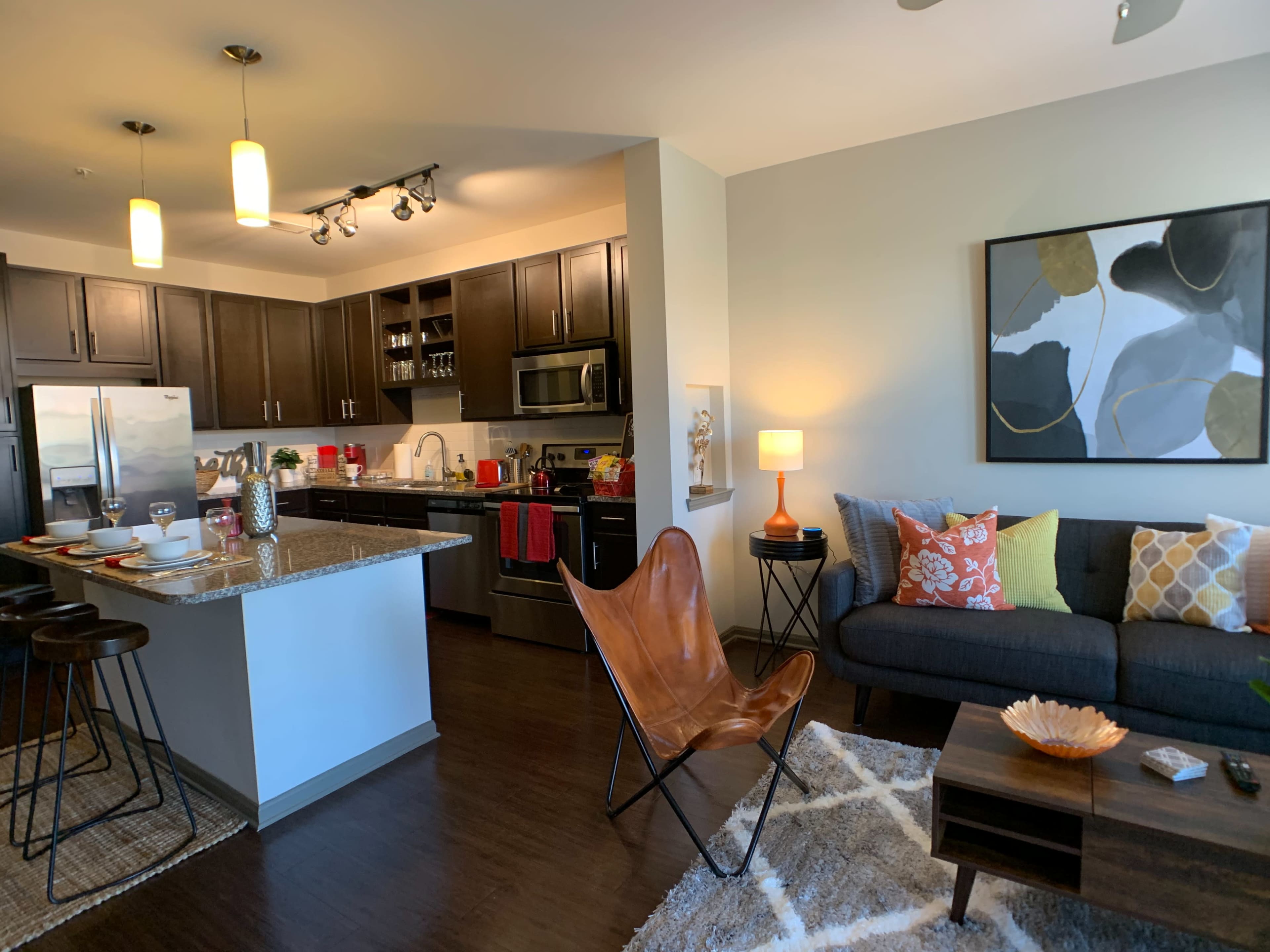 A modern kitchen and living room area featuring dark cabinets, a granite countertop, and a brown leather butterfly chair beside a gray couch adorned with various decorative pillows.