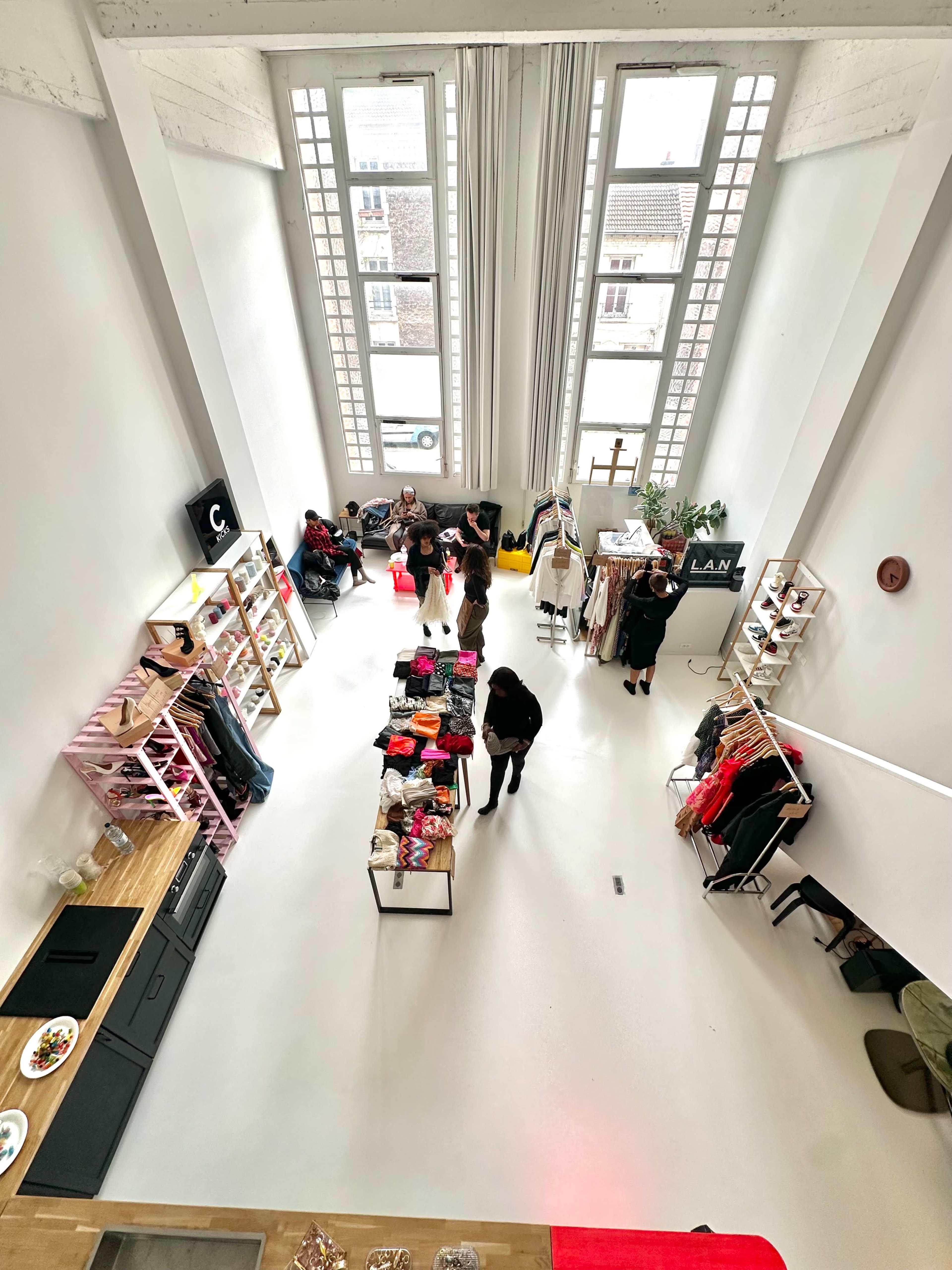 The image shows a spacious clothing retail space with tall windows, various racks of apparel, and a few people browsing and interacting.