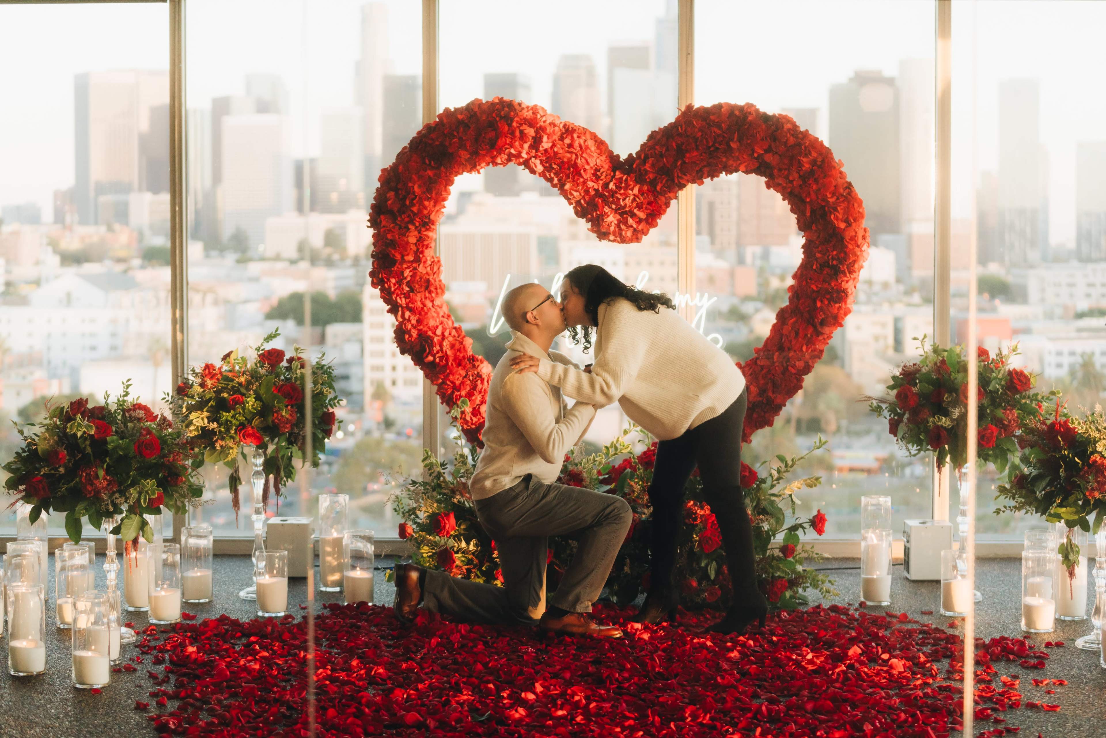 A couple embraces in front of a large heart-shaped floral arrangement made of red roses, set against a city skyline.
