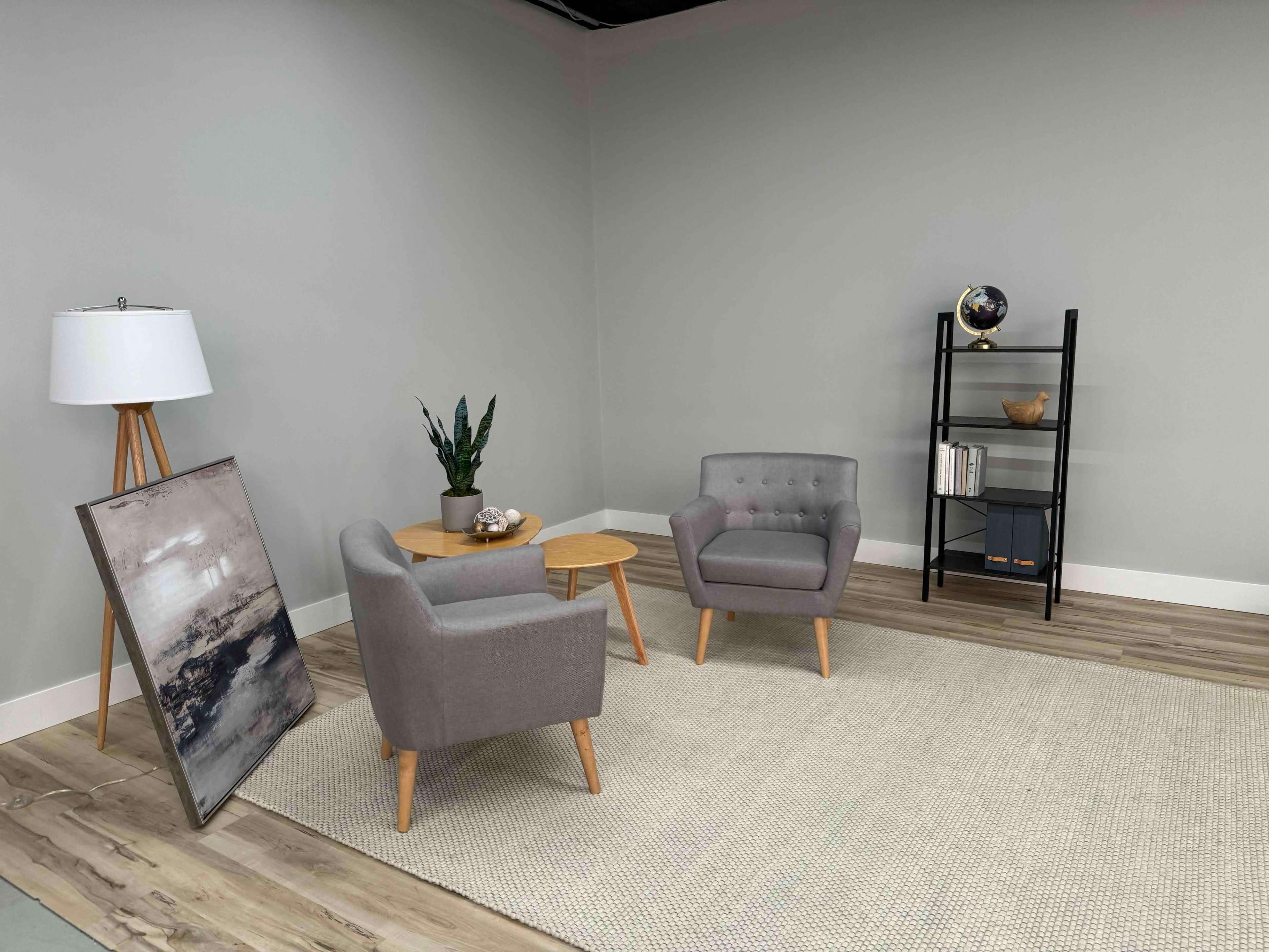 A minimalist living space featuring two gray armchairs, a small wooden table, a floor lamp, a decorative plant, and a black shelving unit against a neutral wall.