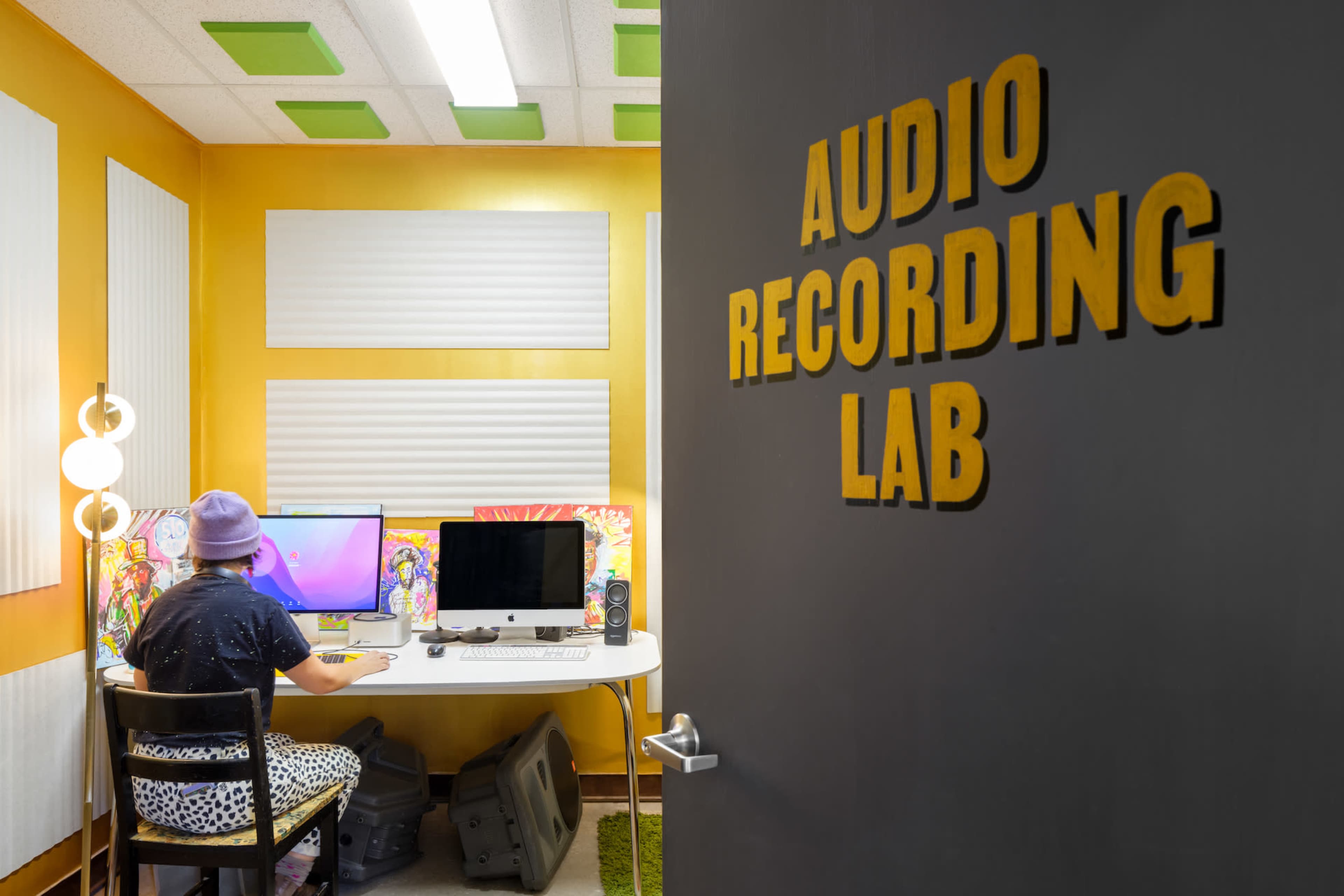 A person sits at a desk with computer monitors in an audio recording lab, illuminated by overhead lights and surrounded by soundproofing panels.