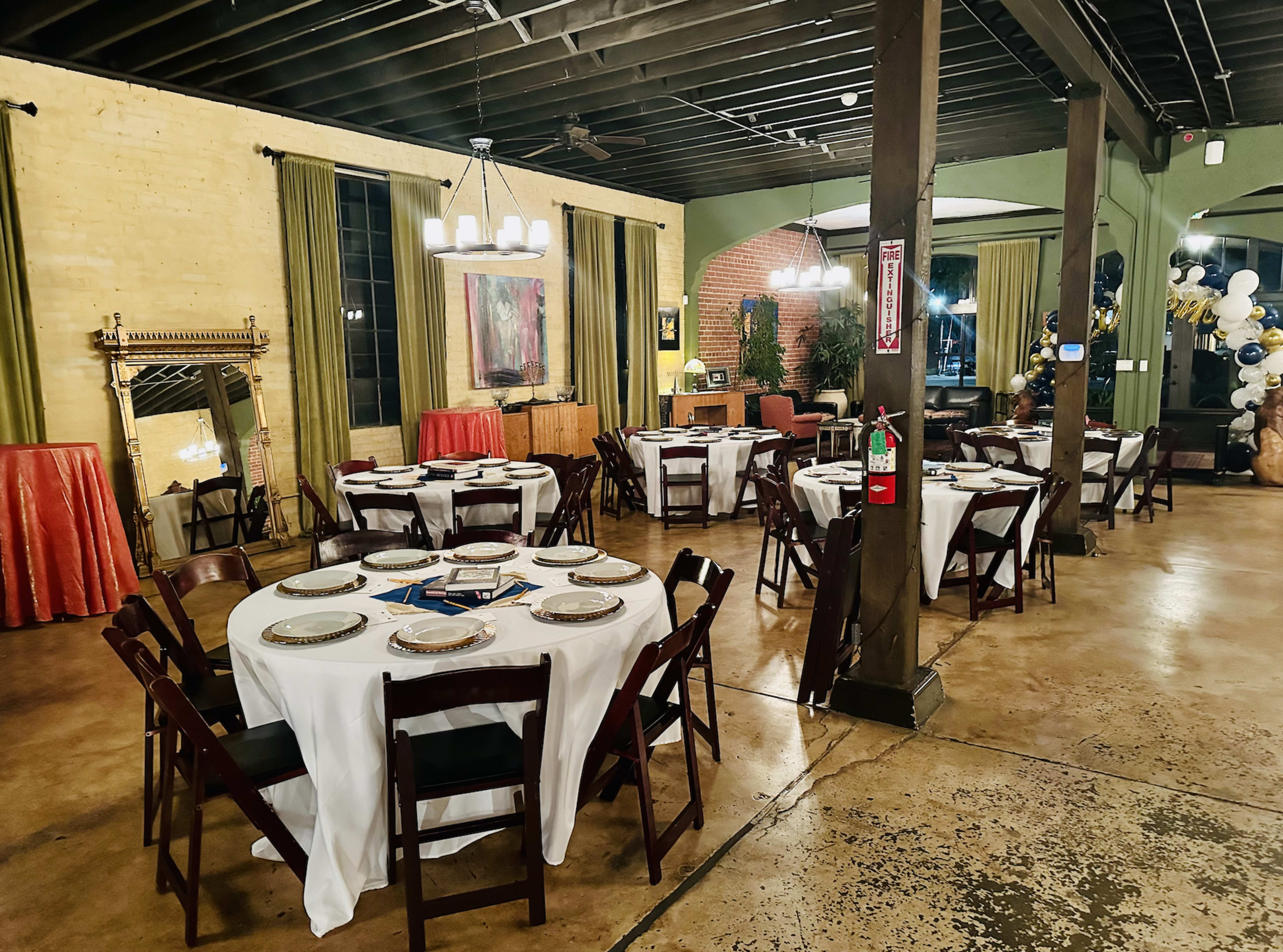 A spacious dining area set with round tables covered in white tablecloths and arranged with plates, located in a room with exposed beams and a mix of brick and painted walls.