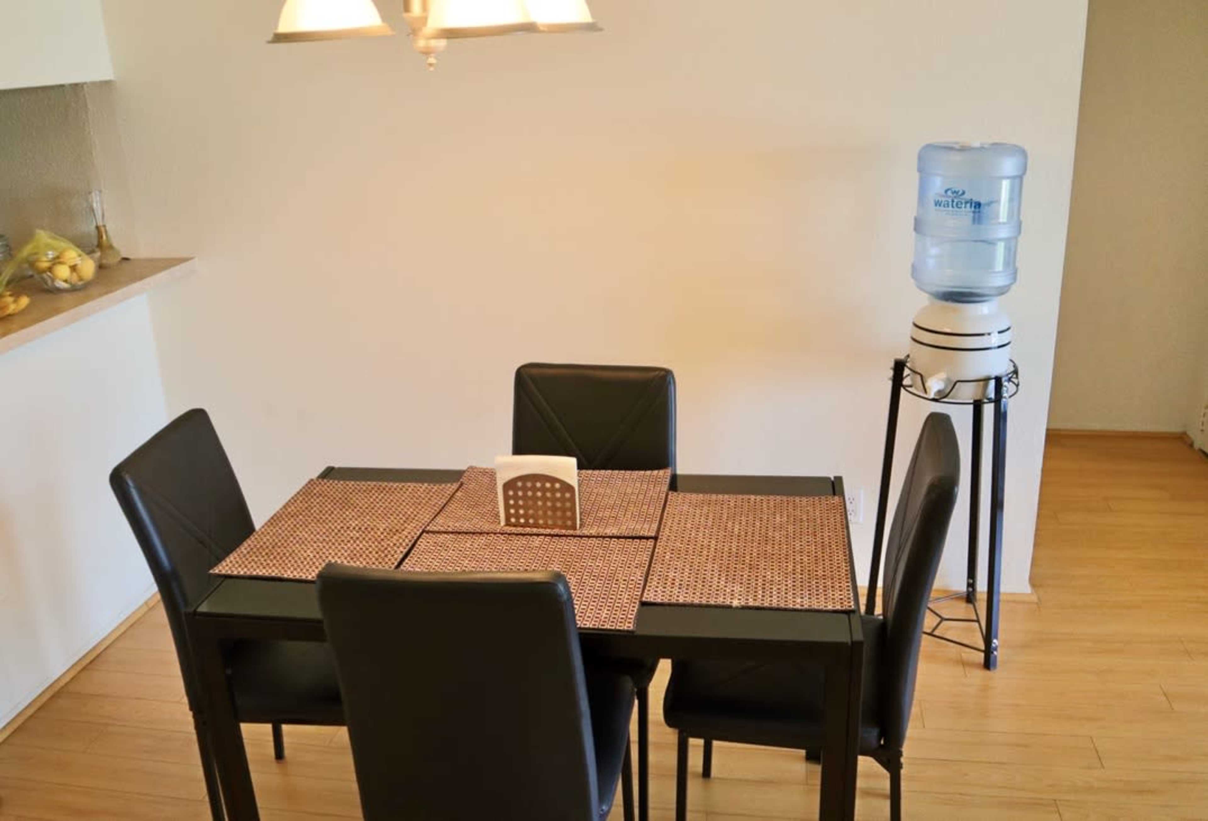 A black dining table with four chairs is set in a room, accompanied by a water cooler positioned on a stand.