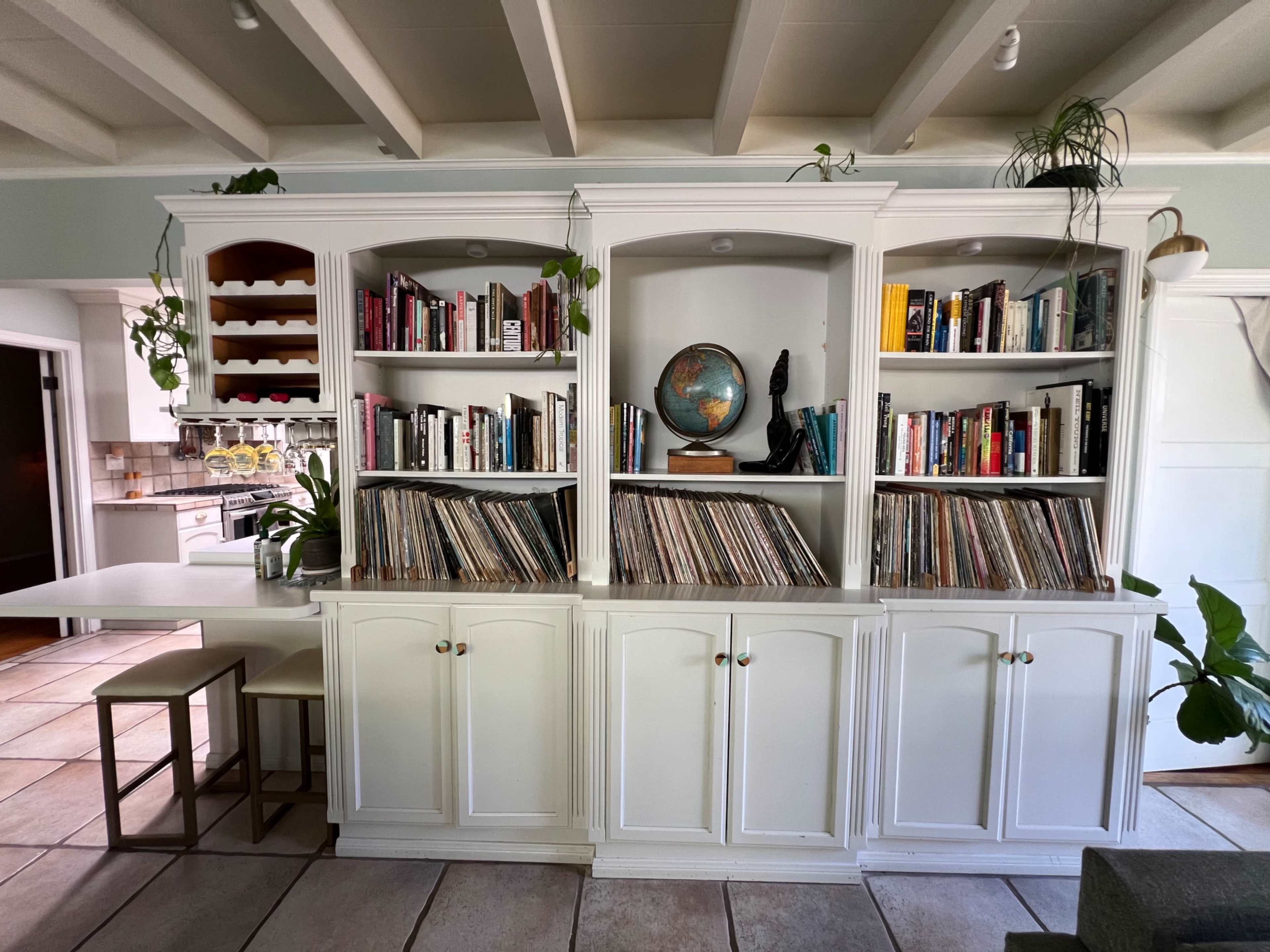 A white bookshelf with several shelves filled with books and records is positioned between a kitchen area and a seating area.