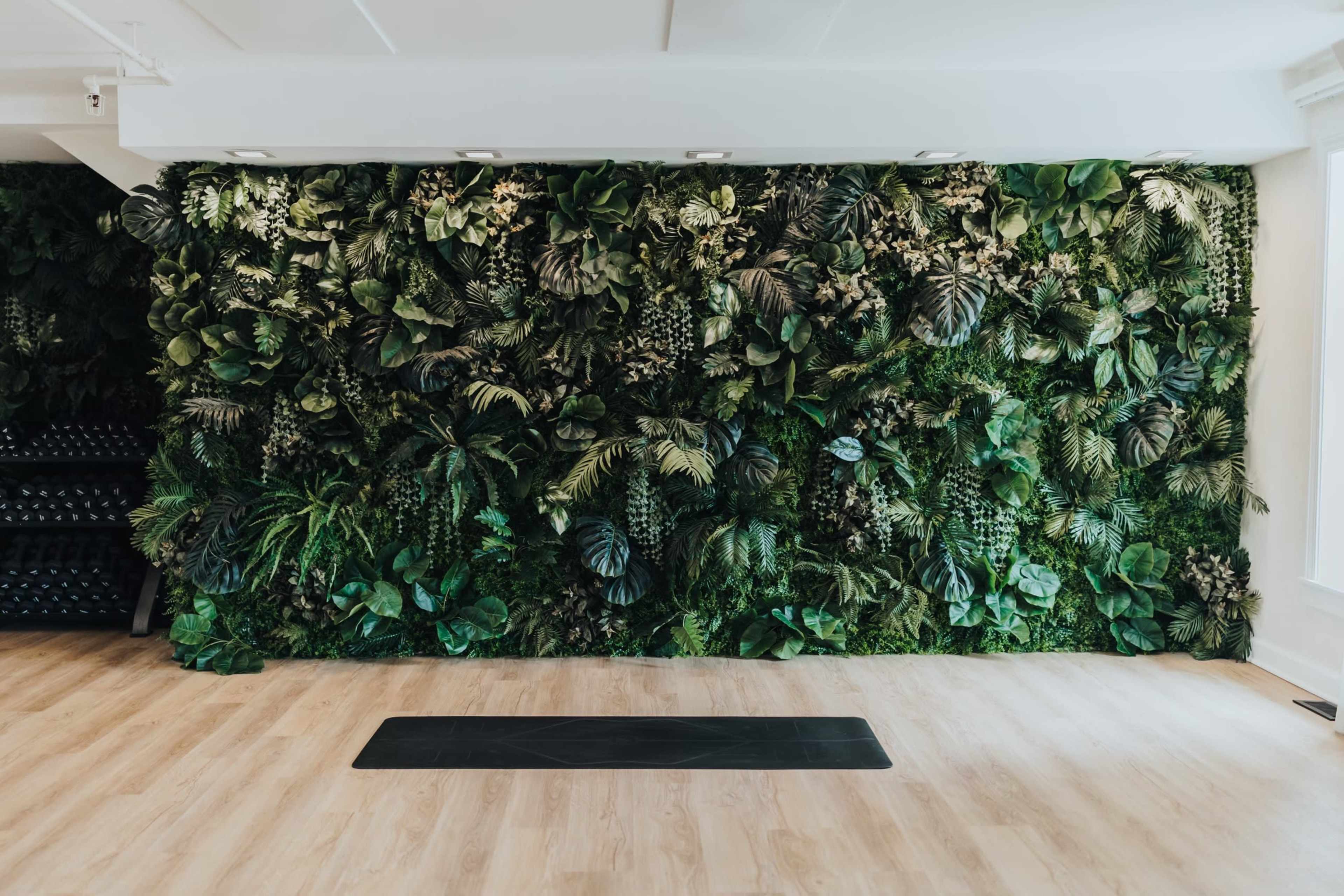 A fitness studio with a textured green wall covered in various artificial plants, complemented by a wooden floor and a yoga mat in front.