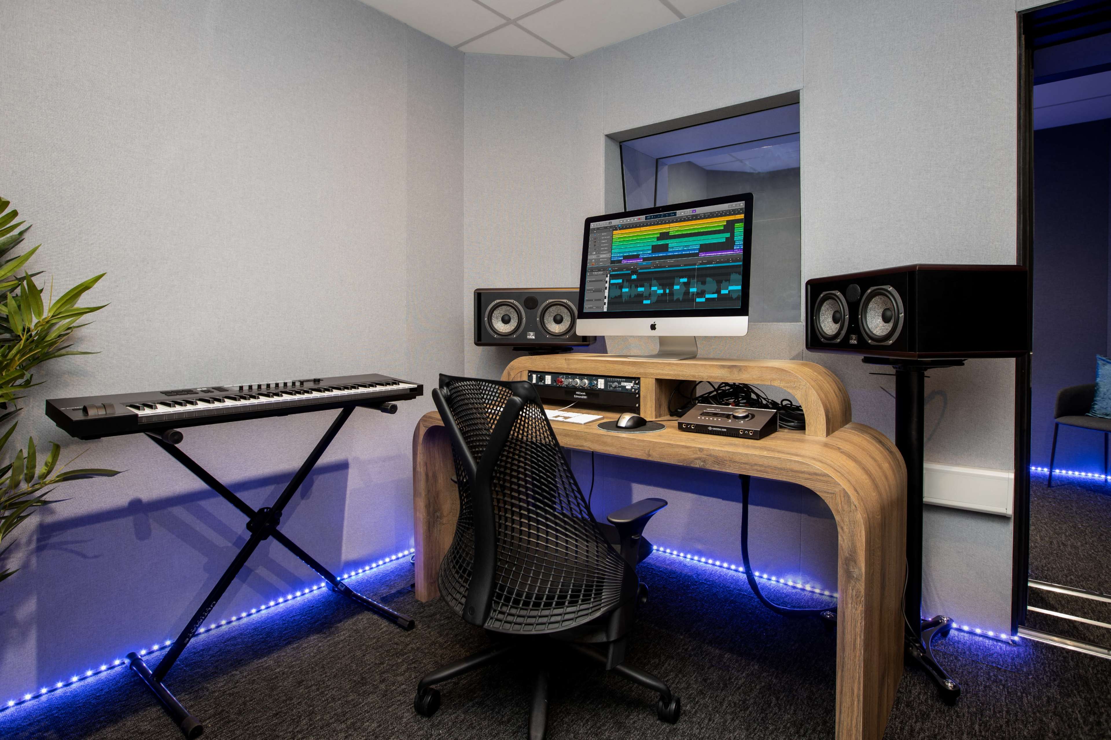 A music production studio featuring a computer desk with a monitor, two speakers, a keyboard, and a black ergonomic chair.
