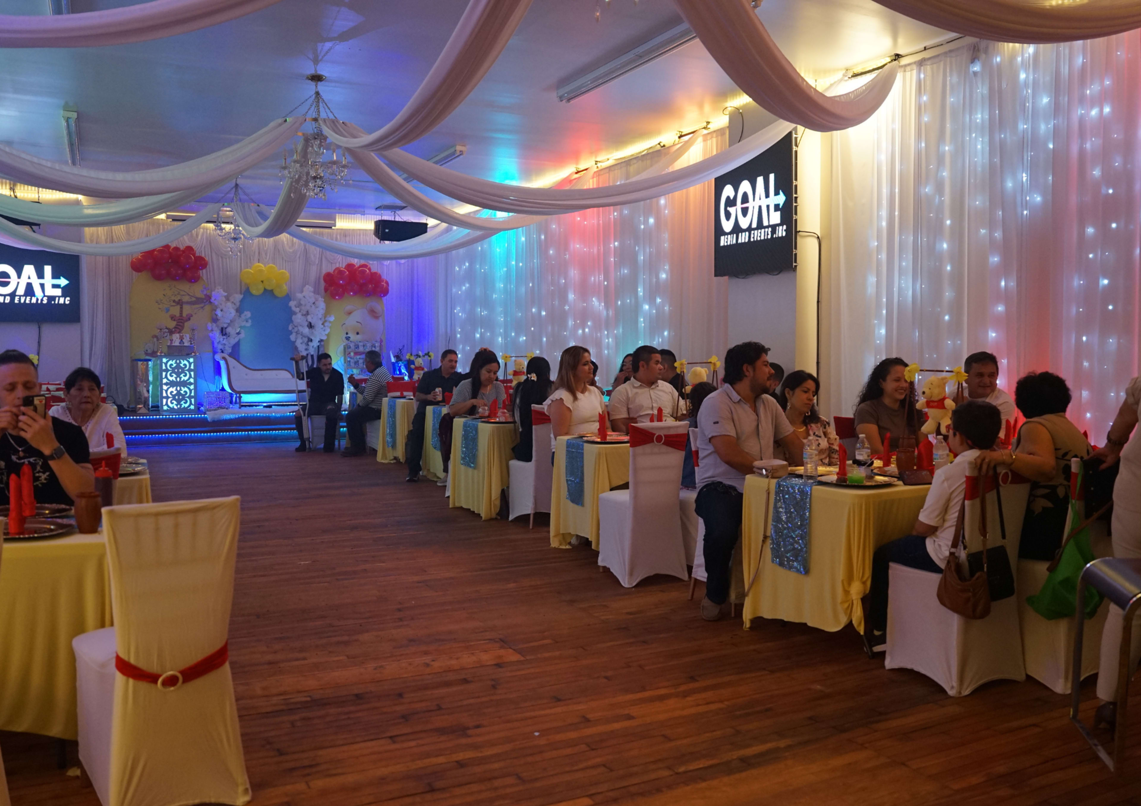 A decorated event space features tables filled with guests enjoying a celebration, accompanied by colorful balloons and lights.