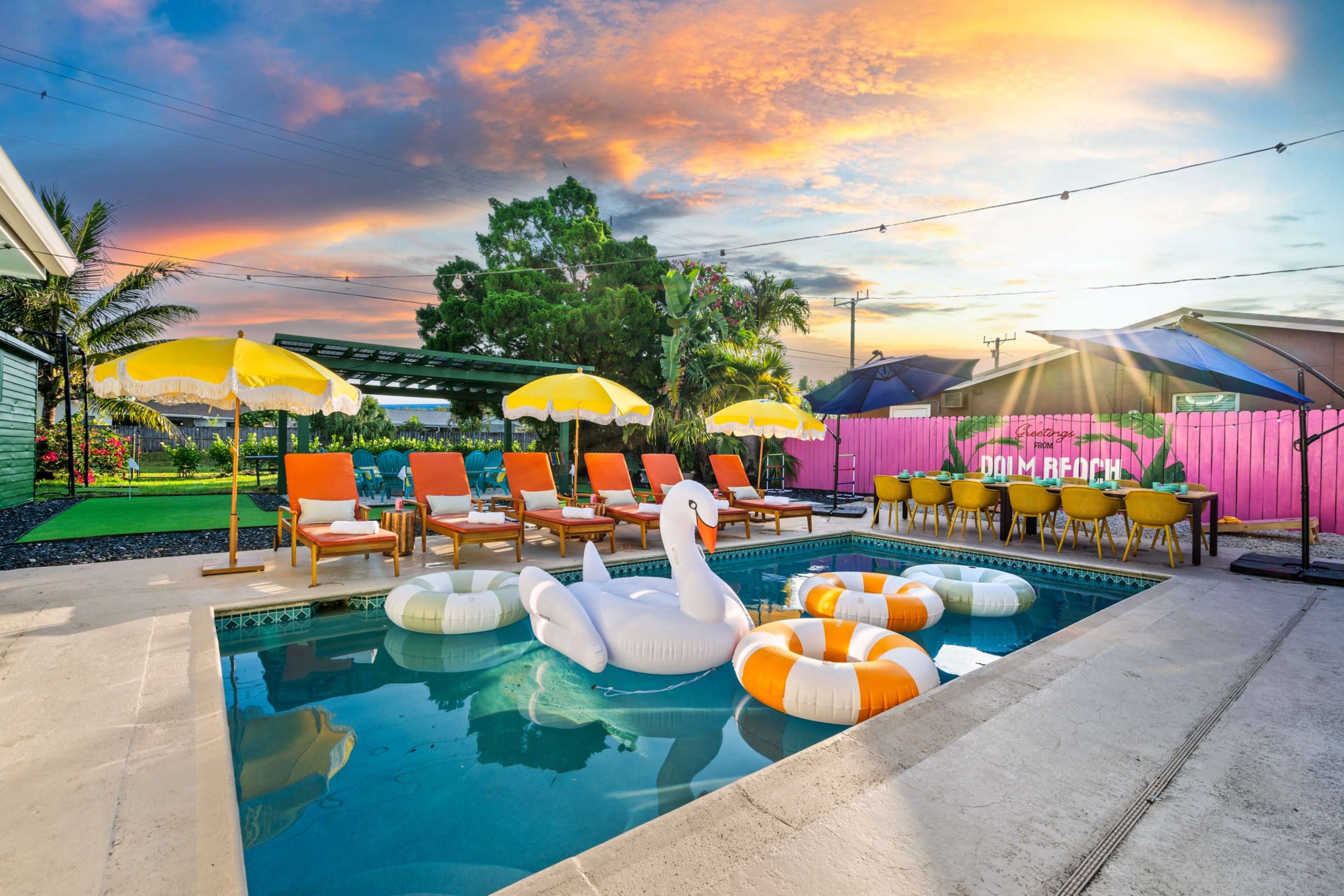 A swimming pool with colorful inflatable floats is surrounded by lounge chairs and yellow umbrellas, set against a vibrant sunset sky.