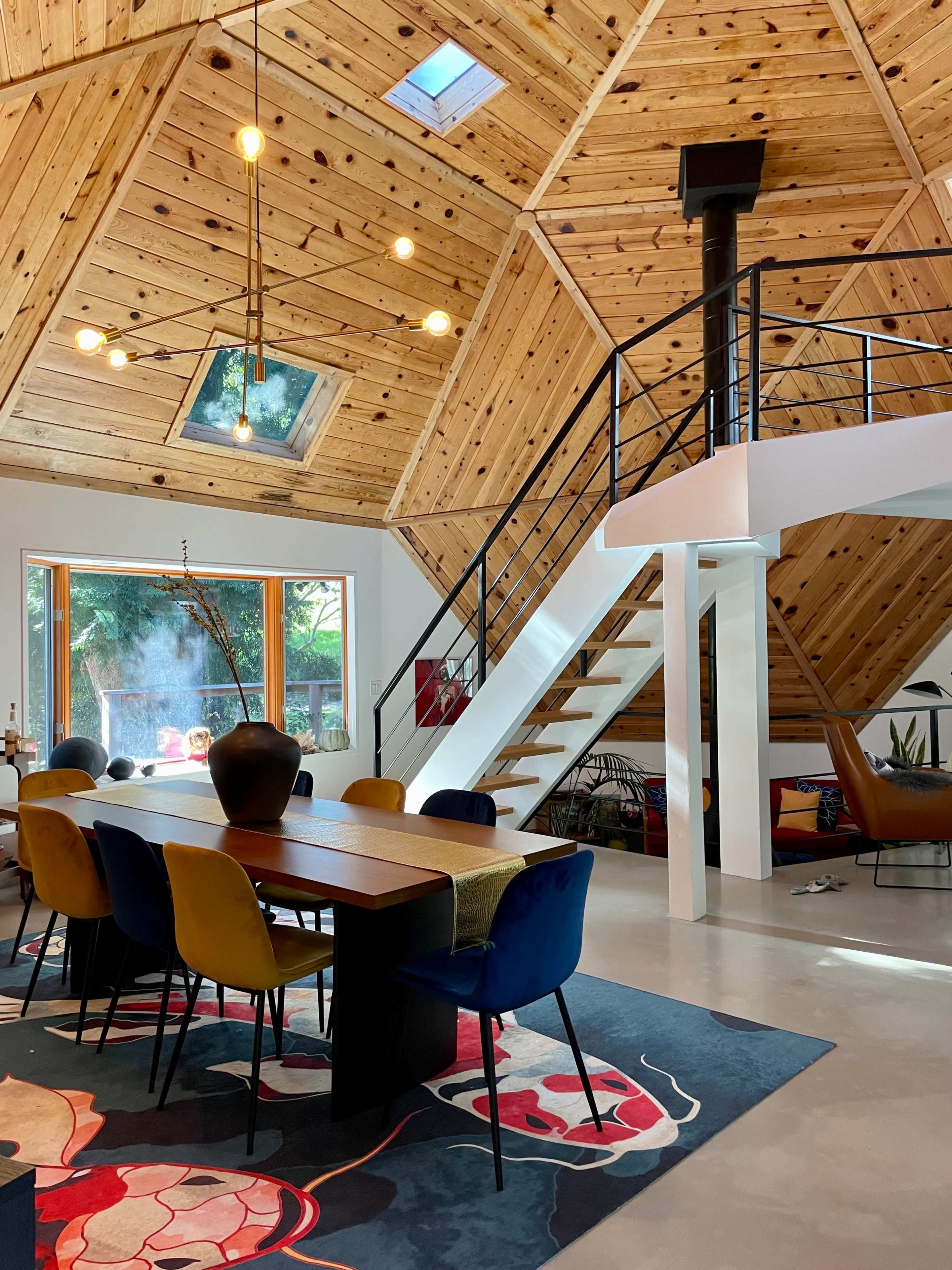 A modern dining area with a large wooden table and colorful chairs, beneath a hexagonal wooden ceiling and a staircase leading to an upper level.