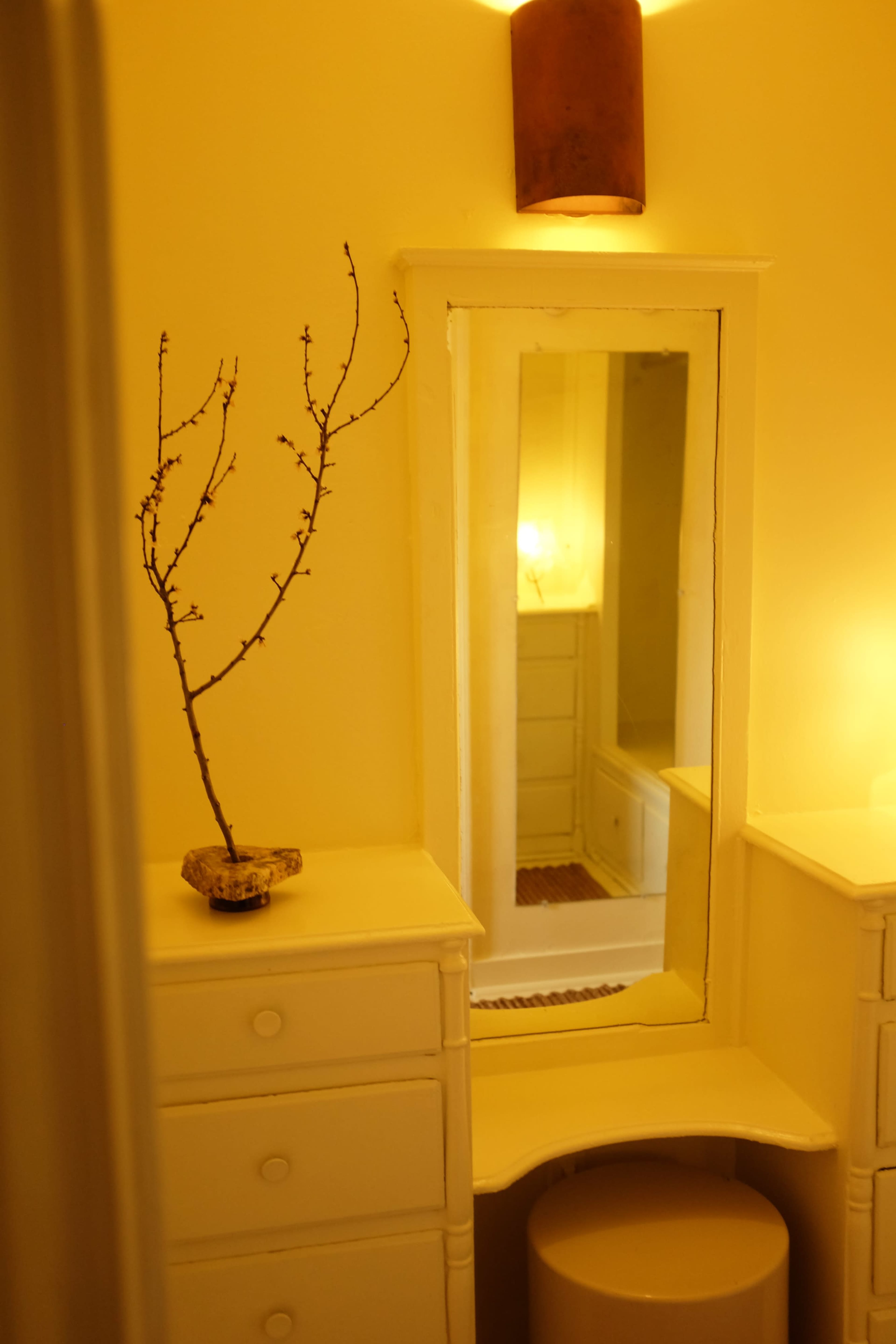 The image shows a softly lit bathroom featuring a round stool, a small white dresser with drawers, and a mirror framed by a simple wall.