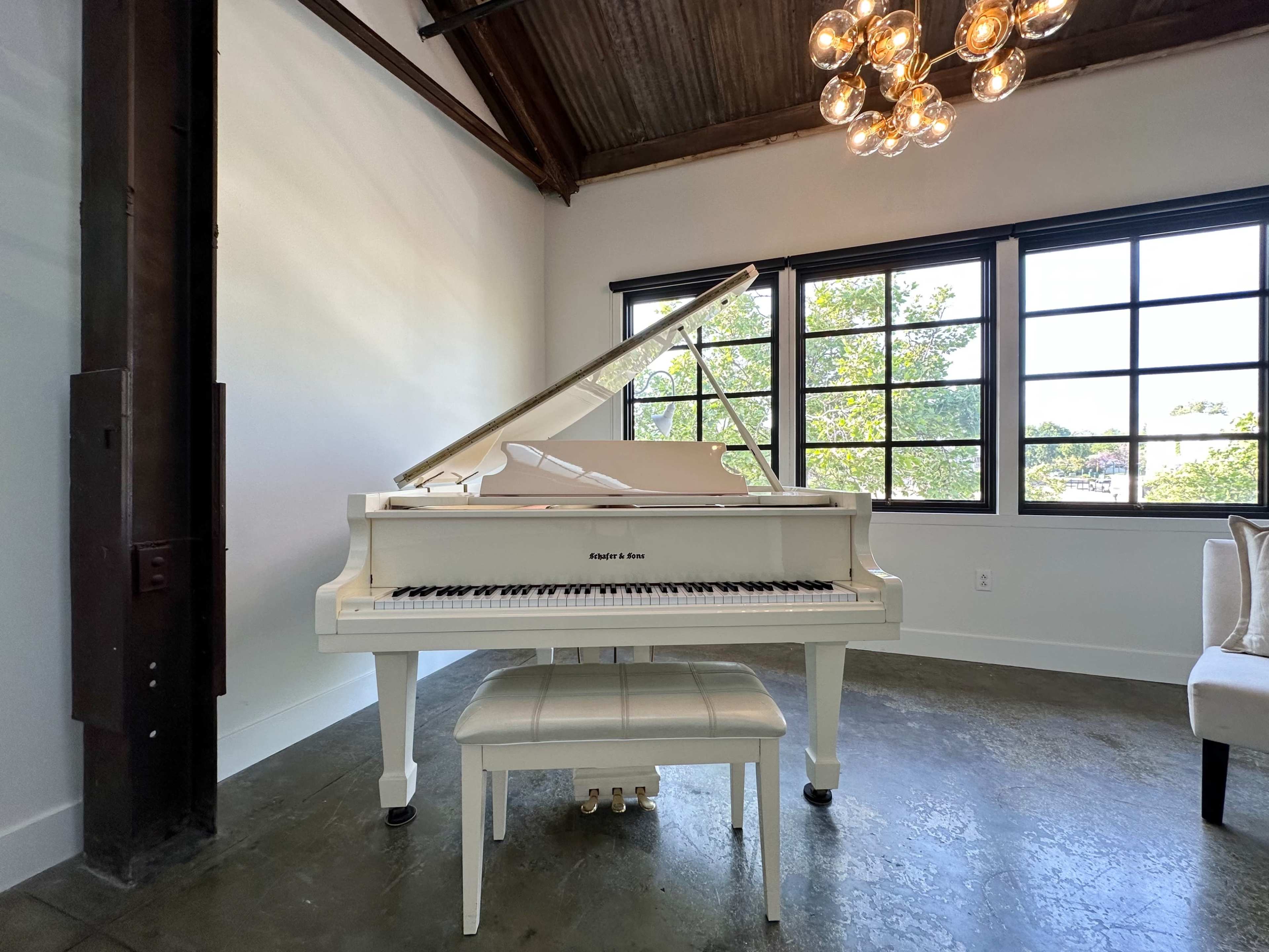 A white grand piano is positioned beside large windows in a well-lit room with industrial-style beams.