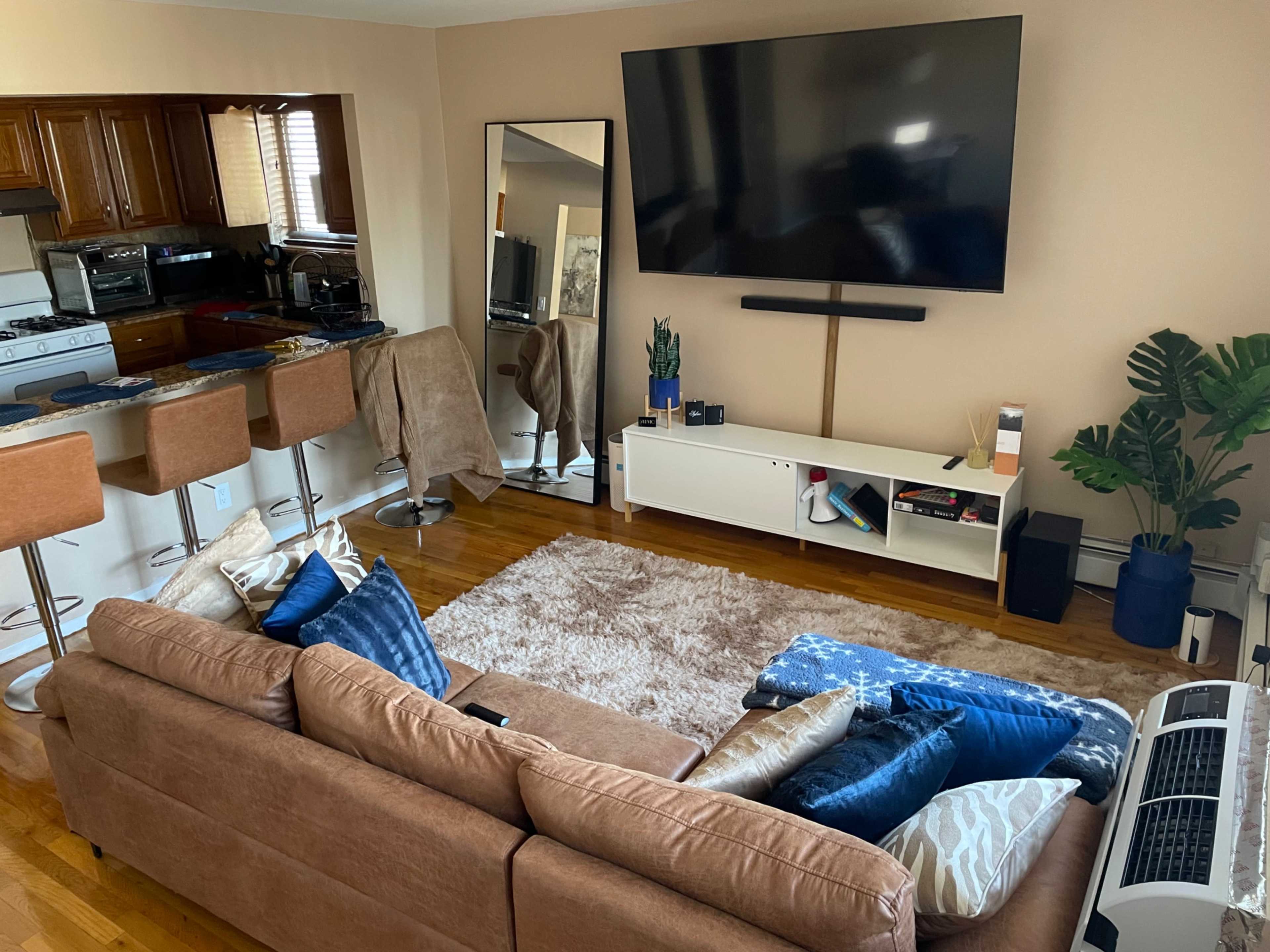 A living room features a brown sectional sofa with various blue cushions, a large wall-mounted TV, a coffee table, and a kitchen area in the background.