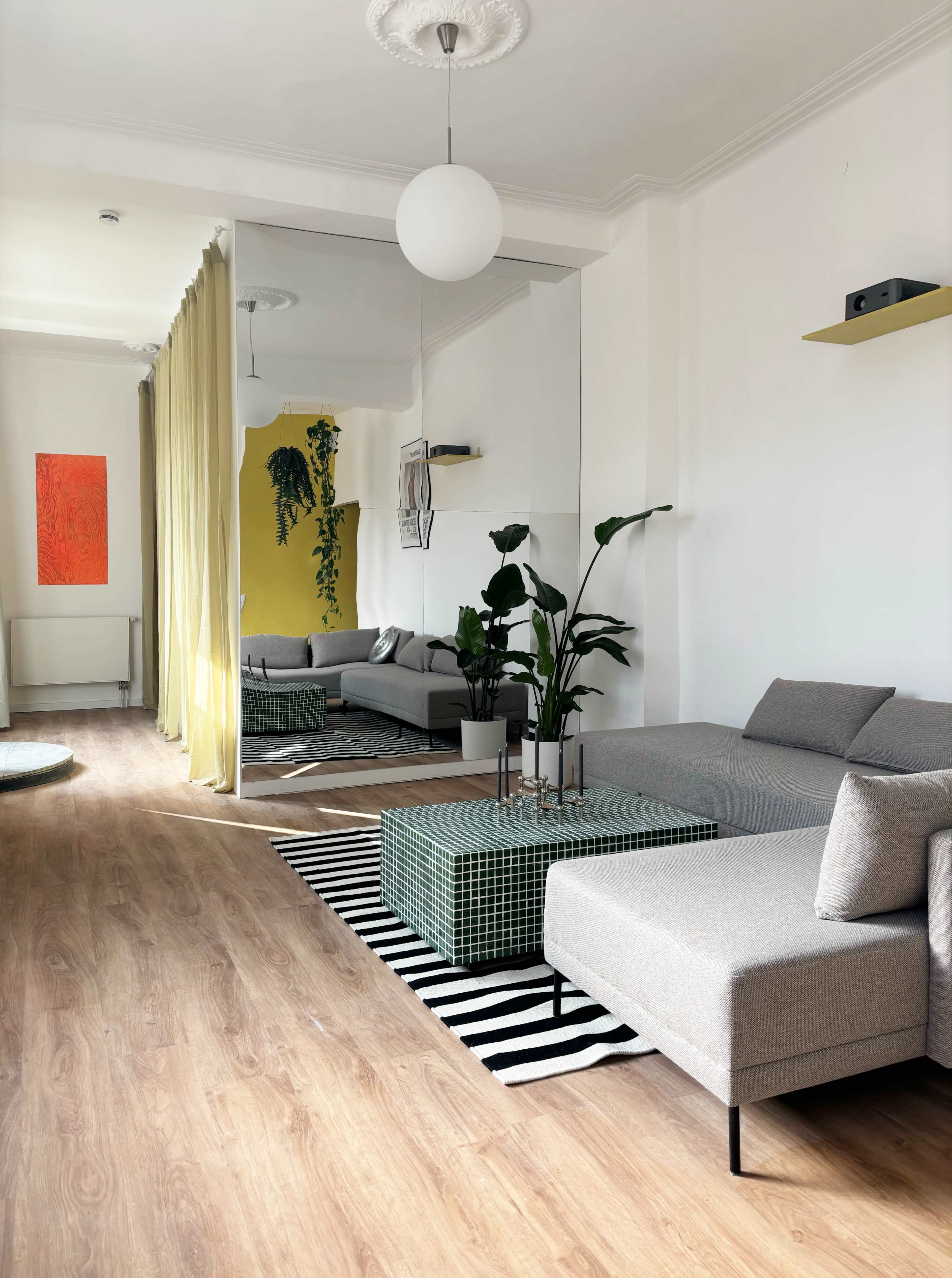 The image shows a modern living room with a gray sectional sofa, a green tiled coffee table, yellow and orange artwork on the walls, and a large mirror reflecting the space.