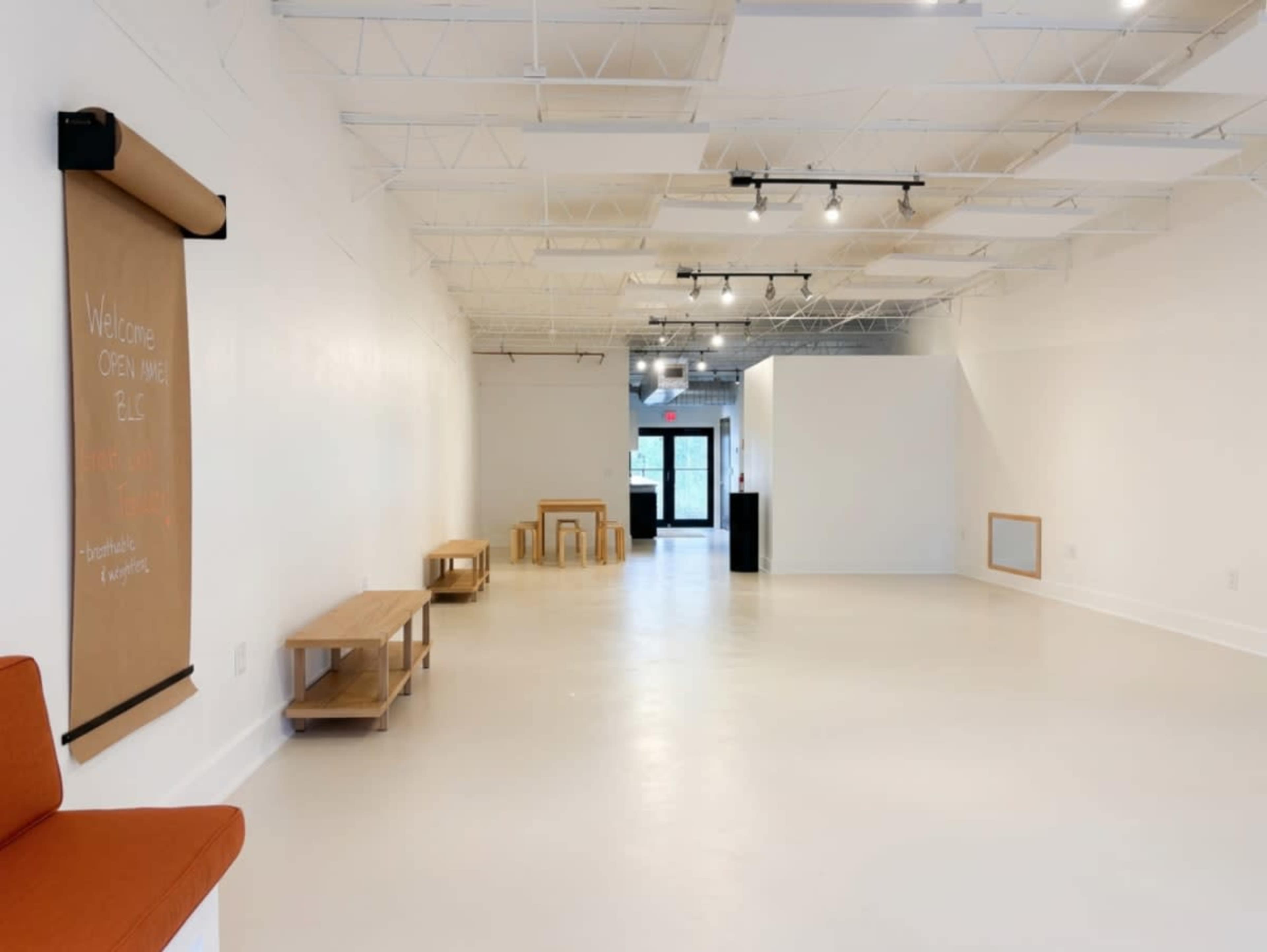 The image shows a spacious, minimalist interior of a gallery or studio with light-colored walls, a roll-up welcome sign, wooden furniture, and ample overhead lighting.