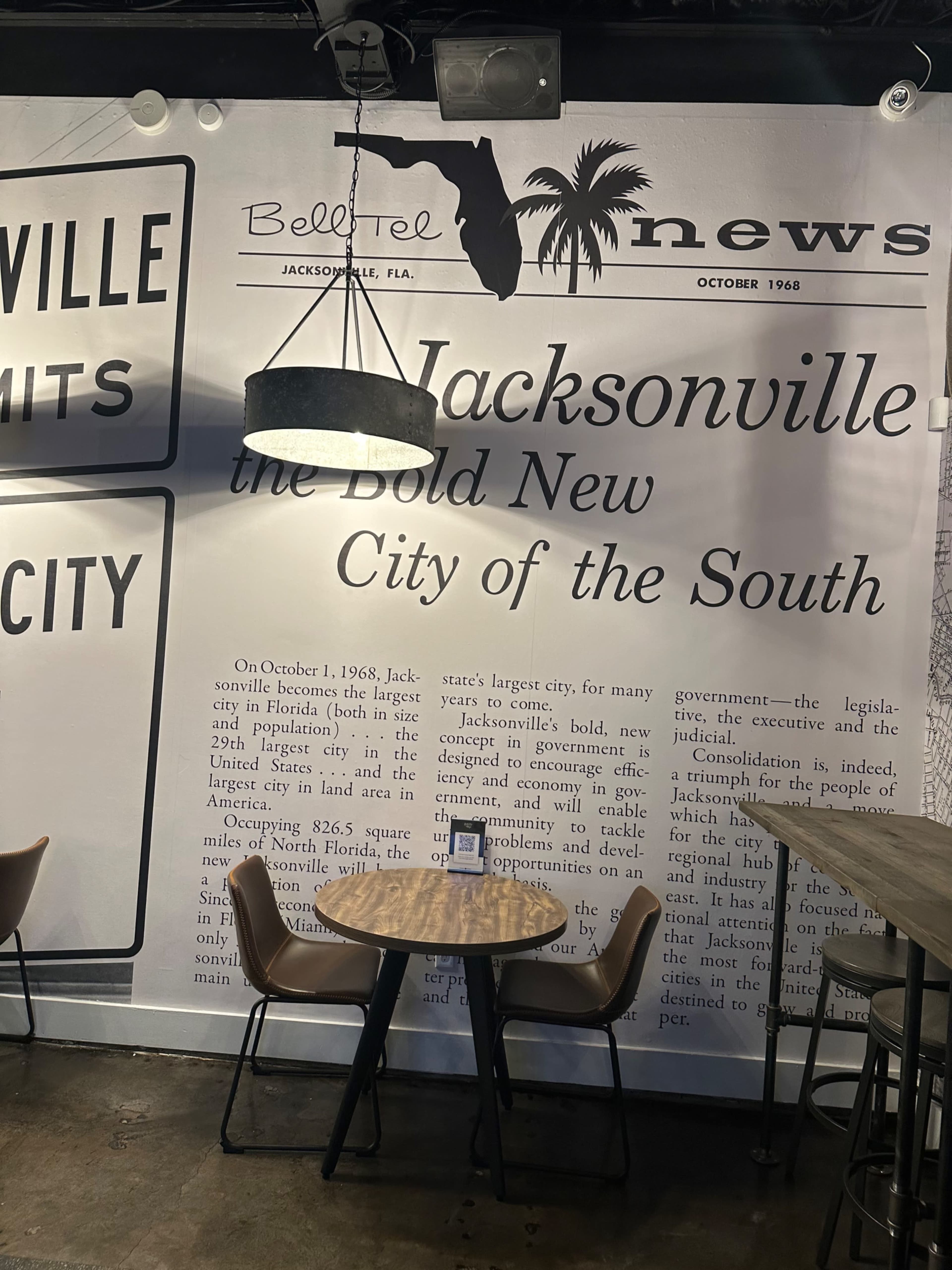 The image shows a cafe setting with a round table and chairs in front of a large wall mural featuring a black and white newspaper-style design, highlighting Jacksonville, Florida, from October 1968.