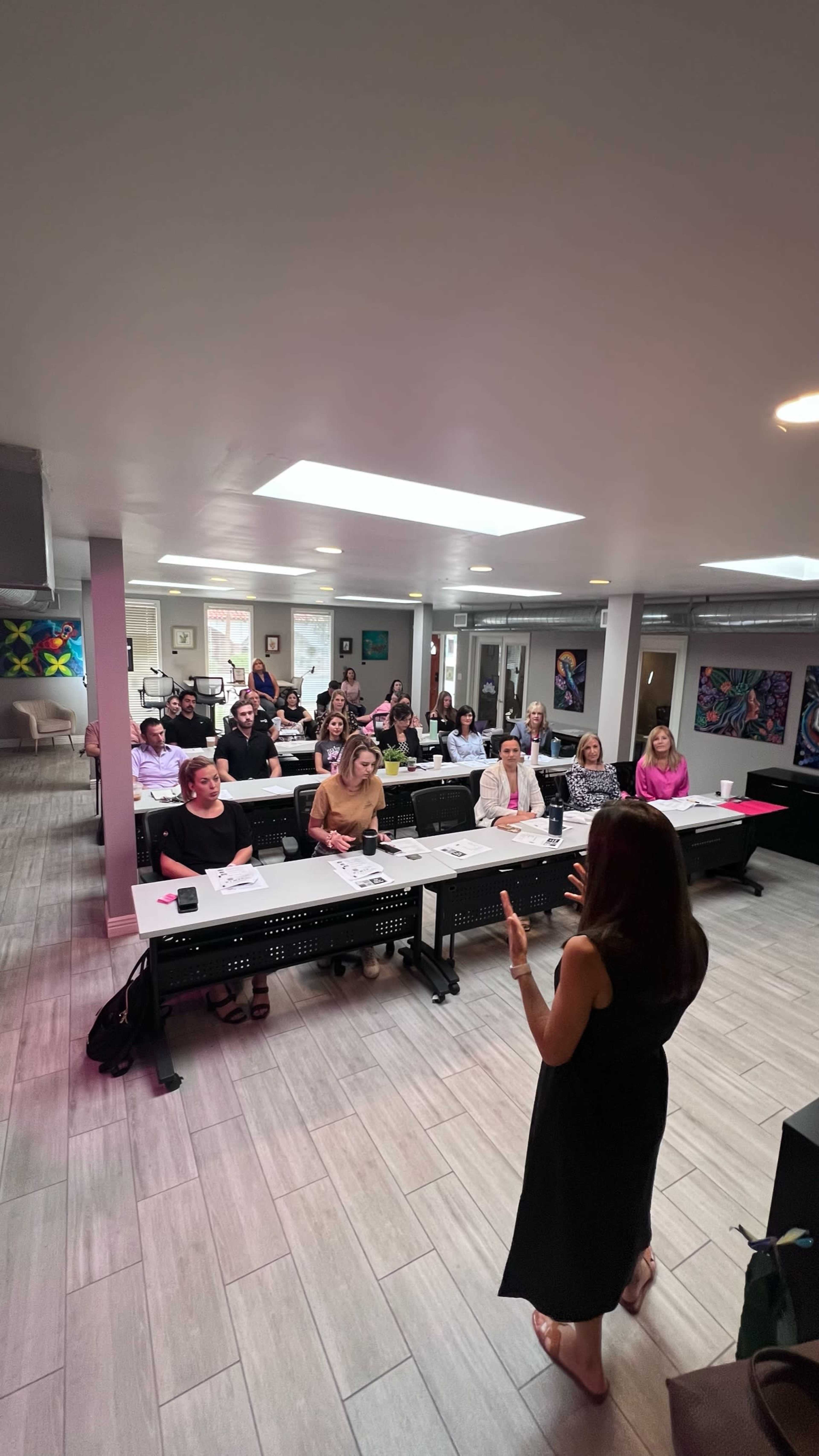 A speaker addresses an audience seated at tables in a well-lit classroom with colorful artwork on the walls.