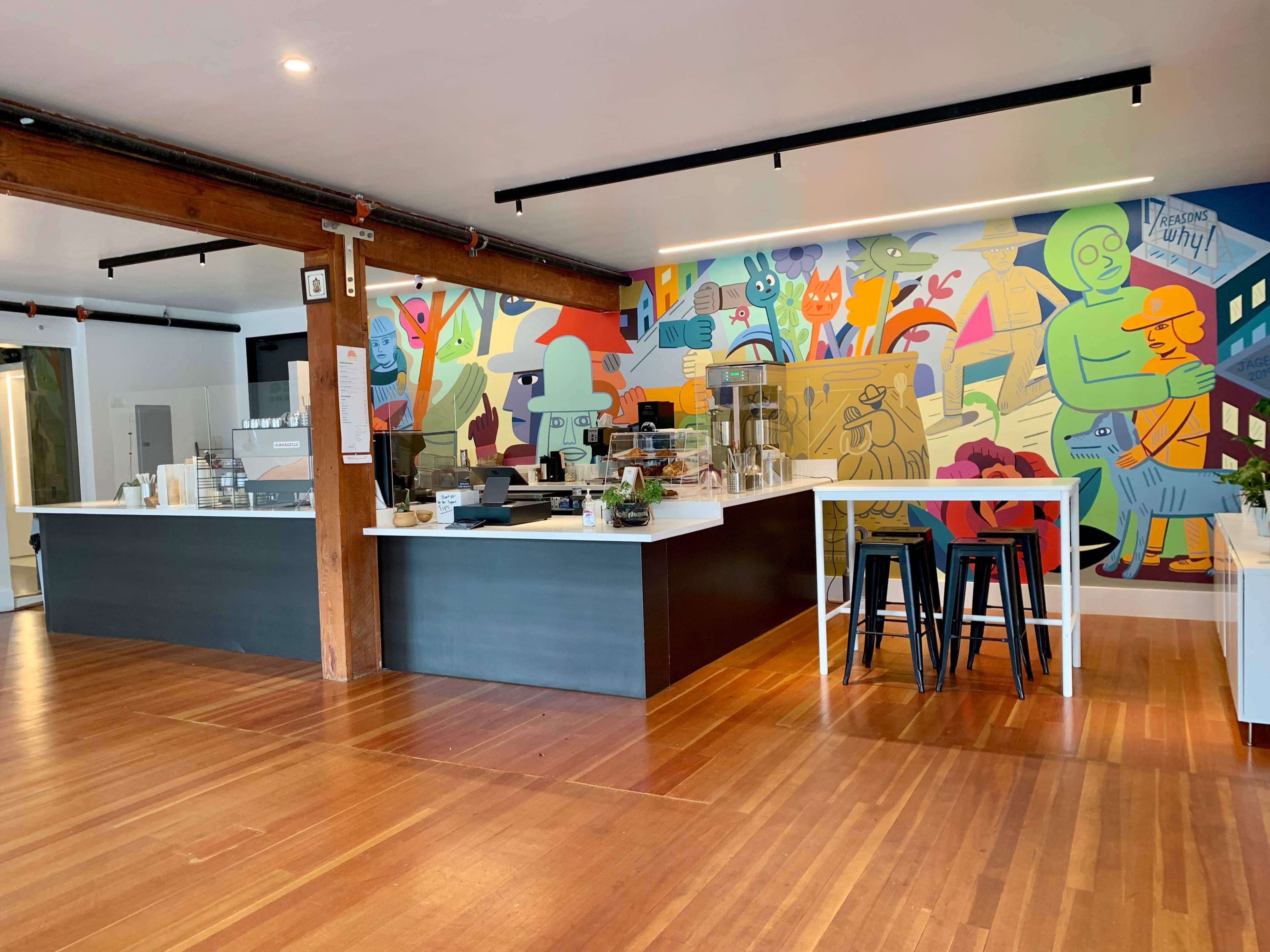 A bright café features a mural of colorful characters on one wall and a service counter with food display cases on the opposite side.