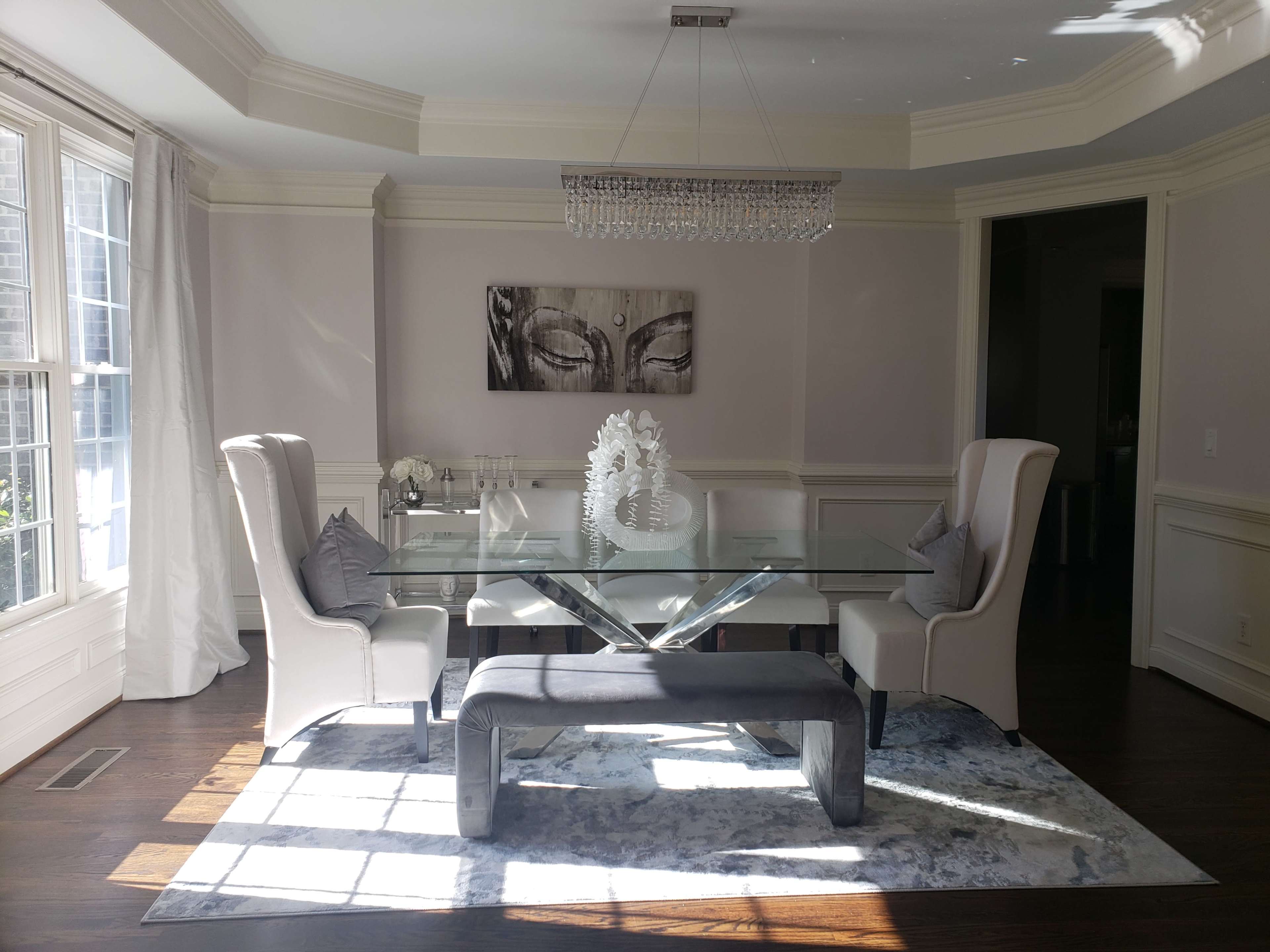 A modern dining room features a glass table surrounded by upholstered chairs, a decorative bench, and a large chandelier above.