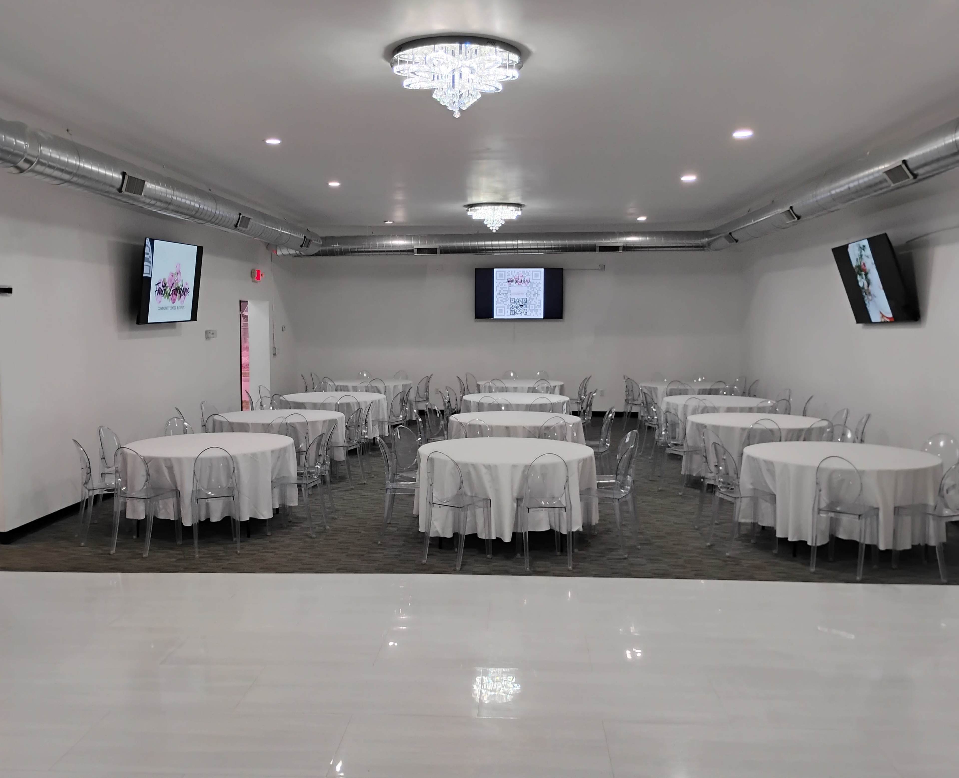 A spacious event room features round tables with white tablecloths and transparent chairs, illuminated by a chandelier, with two large screens on the walls.