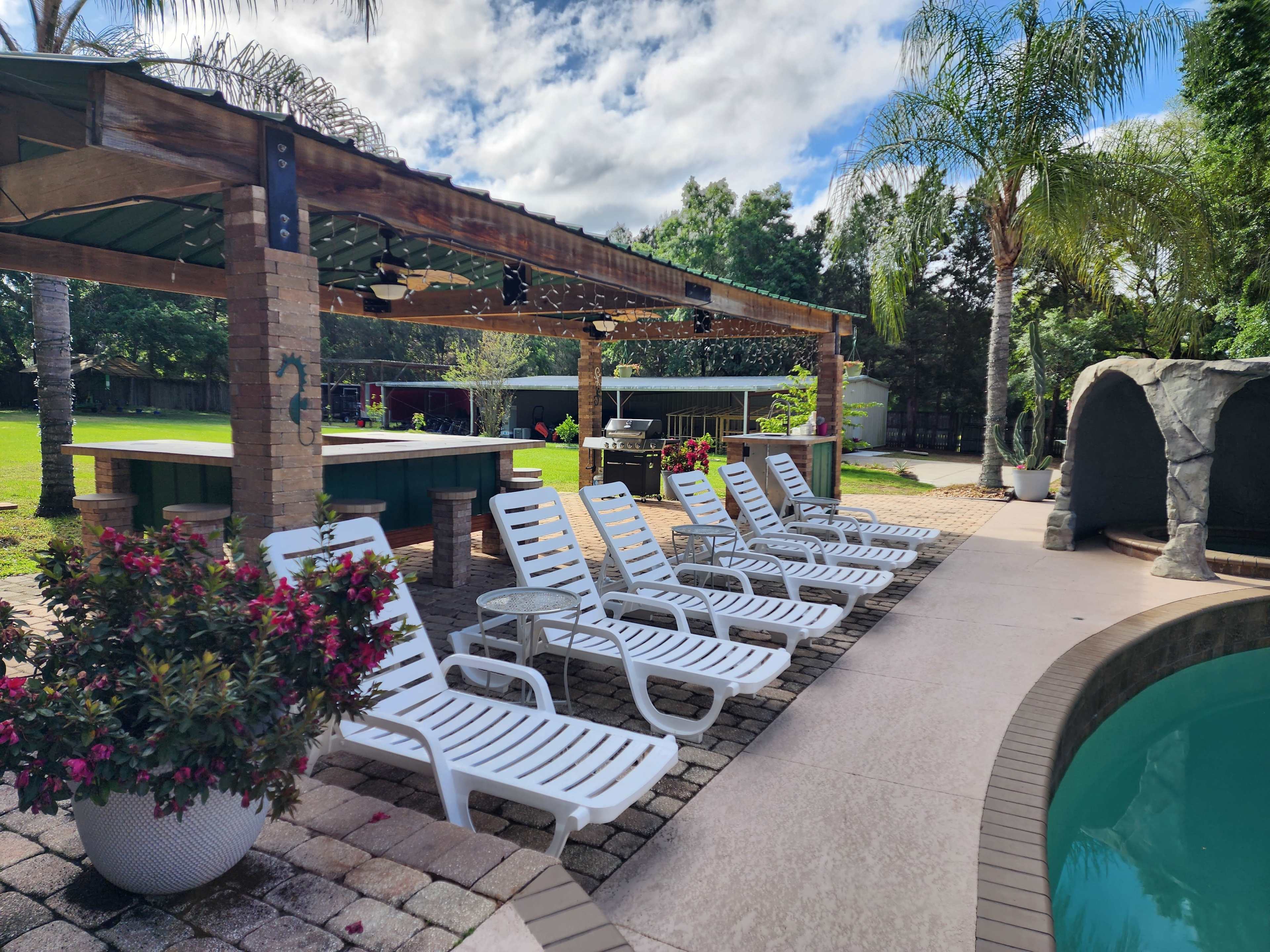 A row of white lounge chairs is arranged beside a swimming pool in a landscaped backyard with a gazebo and greenery.