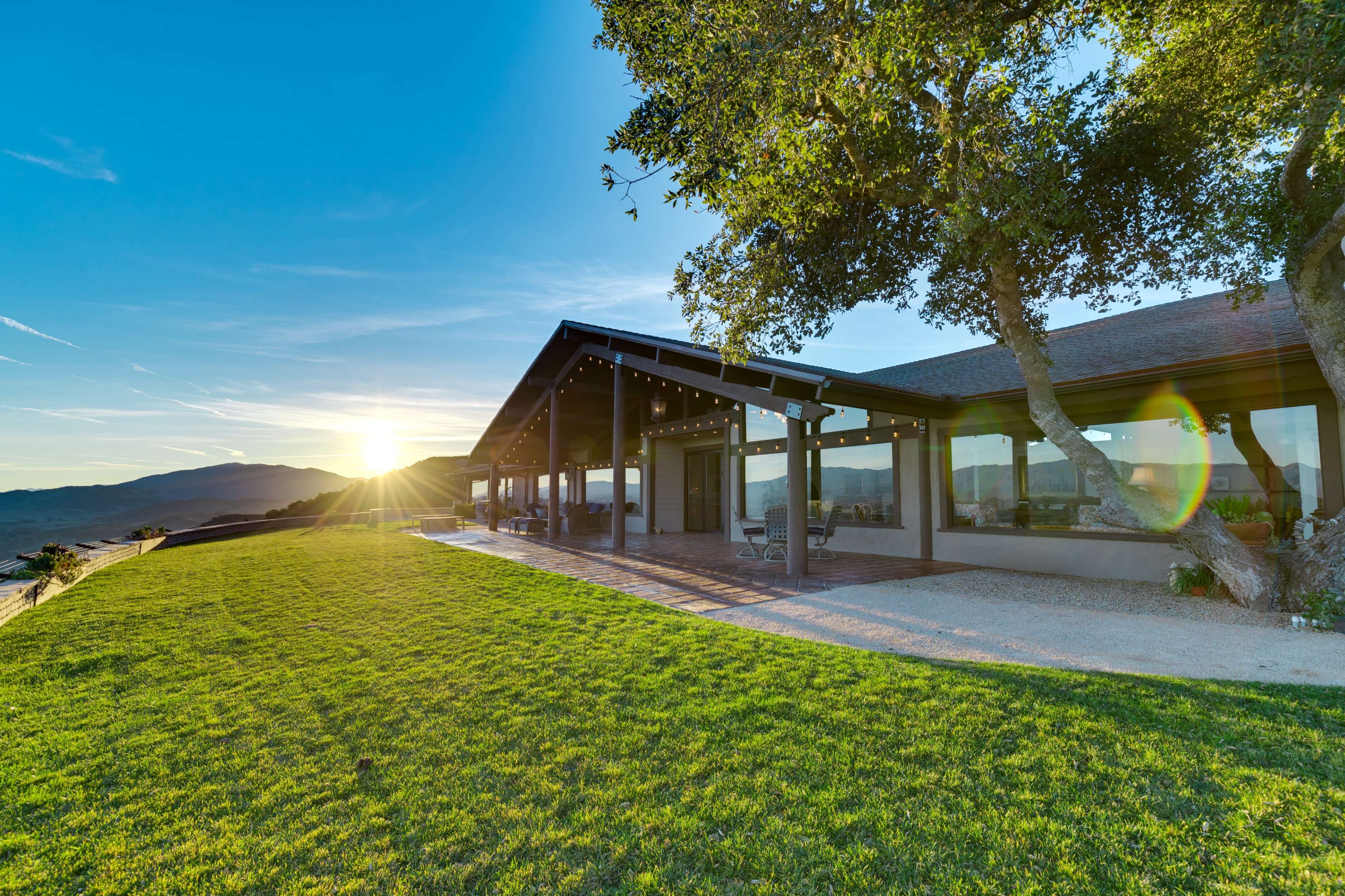 A modern house with large windows is set on a grassy hillside, overlooking distant mountains as the sun sets.