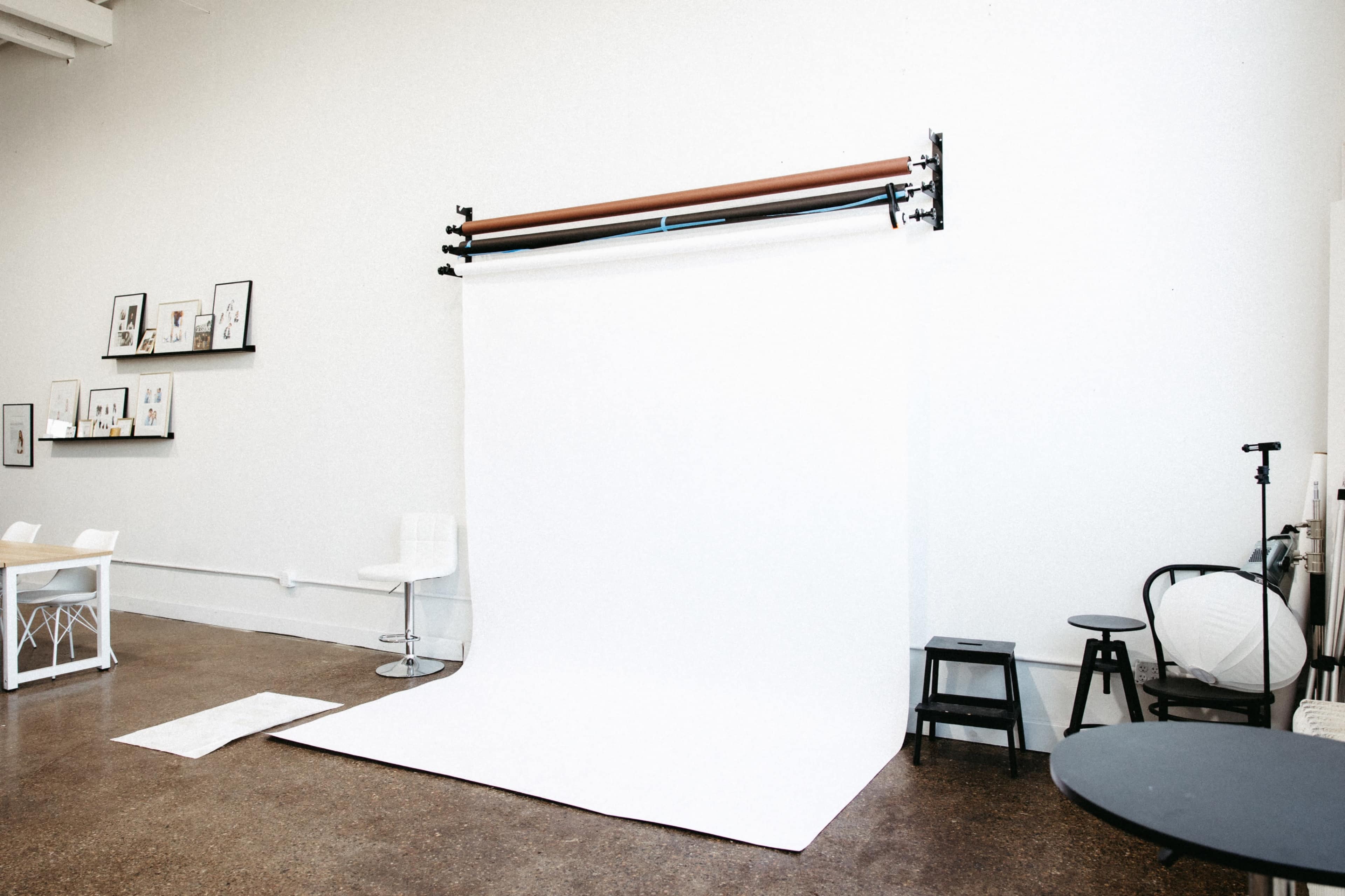 A photography studio features a blank white backdrop with wooden support rods, surrounded by minimalist furniture and displayed artwork on the walls.
