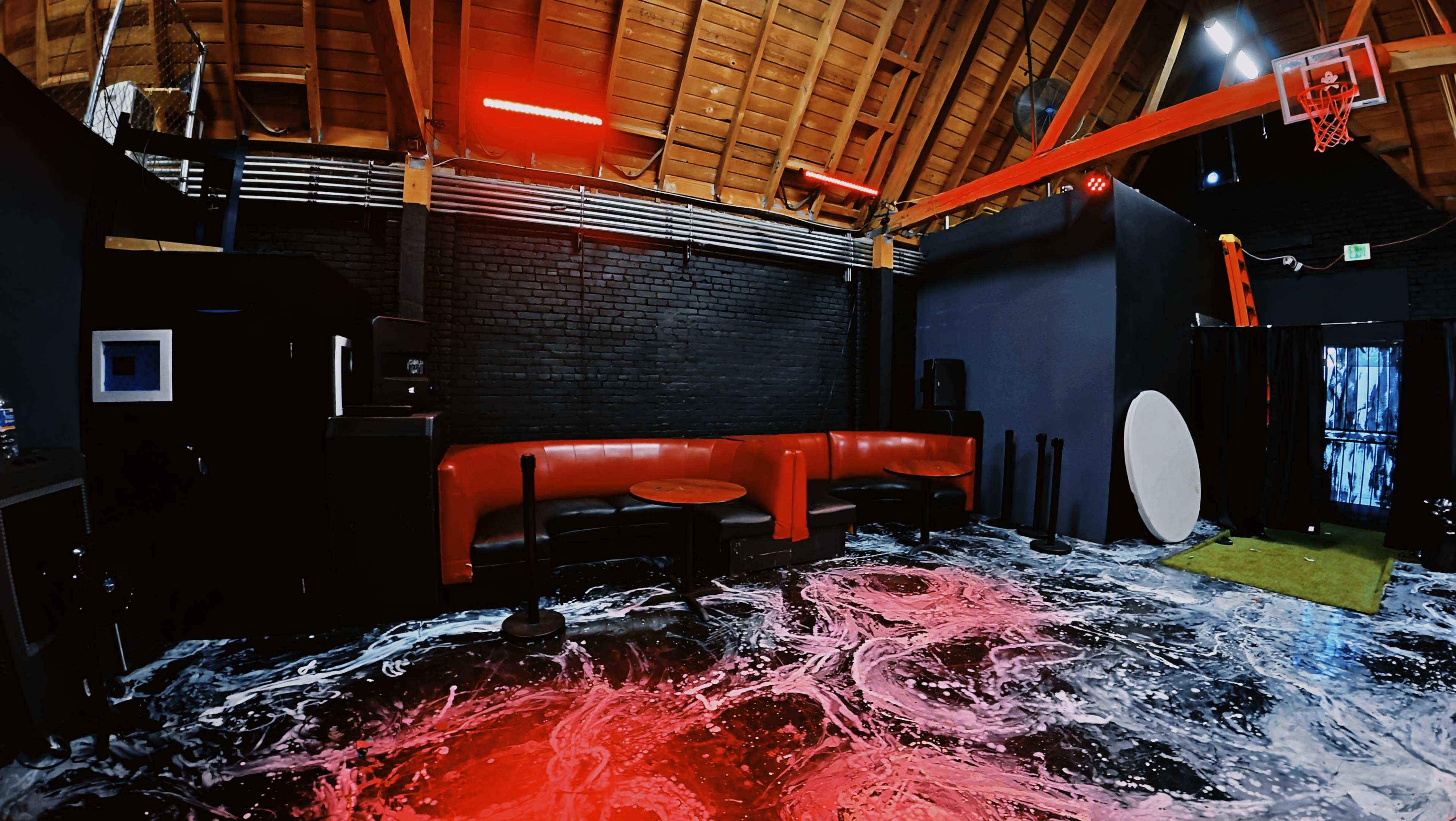 The image shows a spacious indoor area with black walls, a red seating area, and a basketball hoop, featuring a colorful floor design.