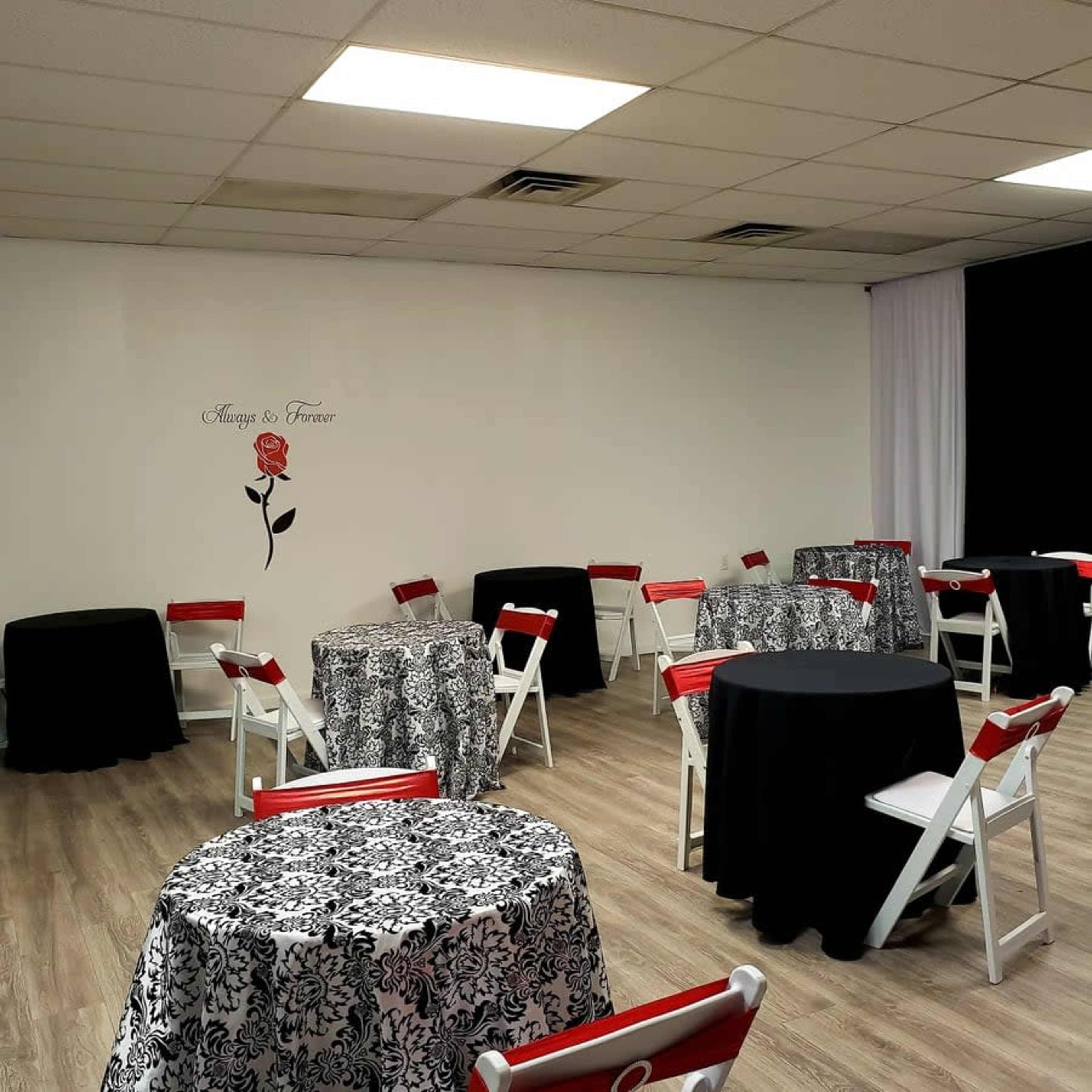 A room set up for an event with several round tables covered in black and white floral tablecloths and decorated with red chairs.