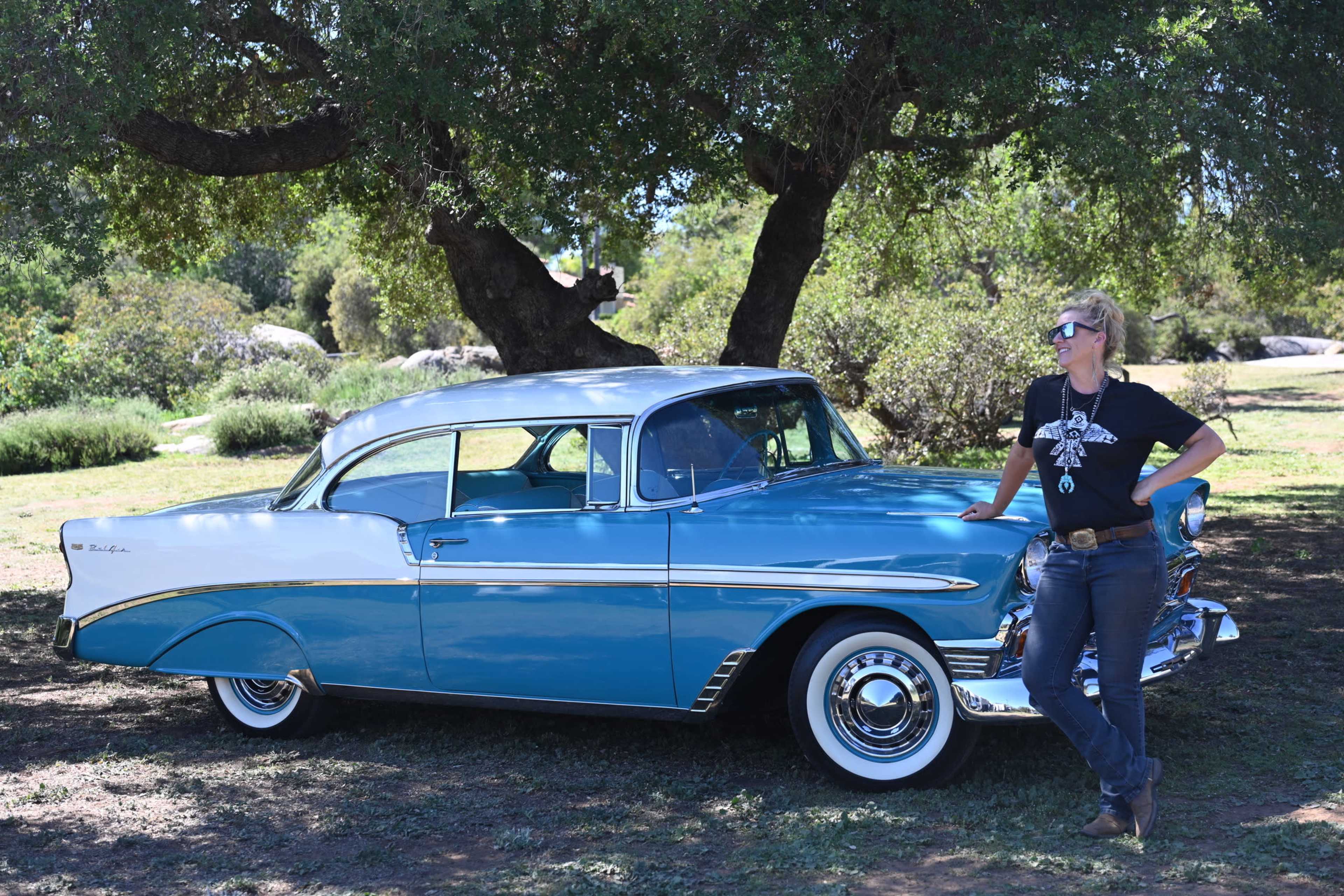 A woman in a black t-shirt stands beside a vintage blue and white car under a large tree.