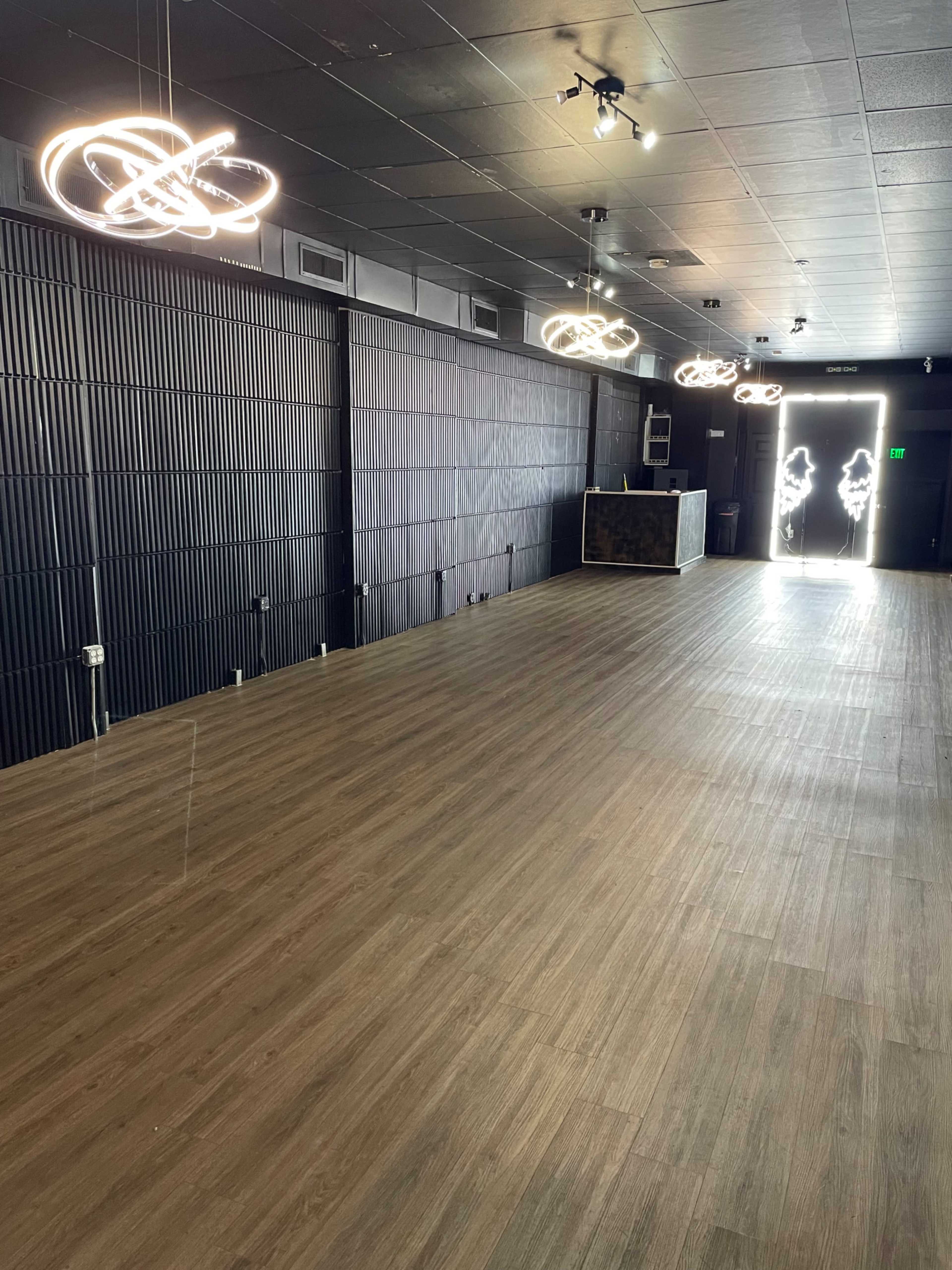 The image shows an empty room with wooden flooring, black walls, modern light fixtures, and an entrance to an outside space illuminated by bright light.