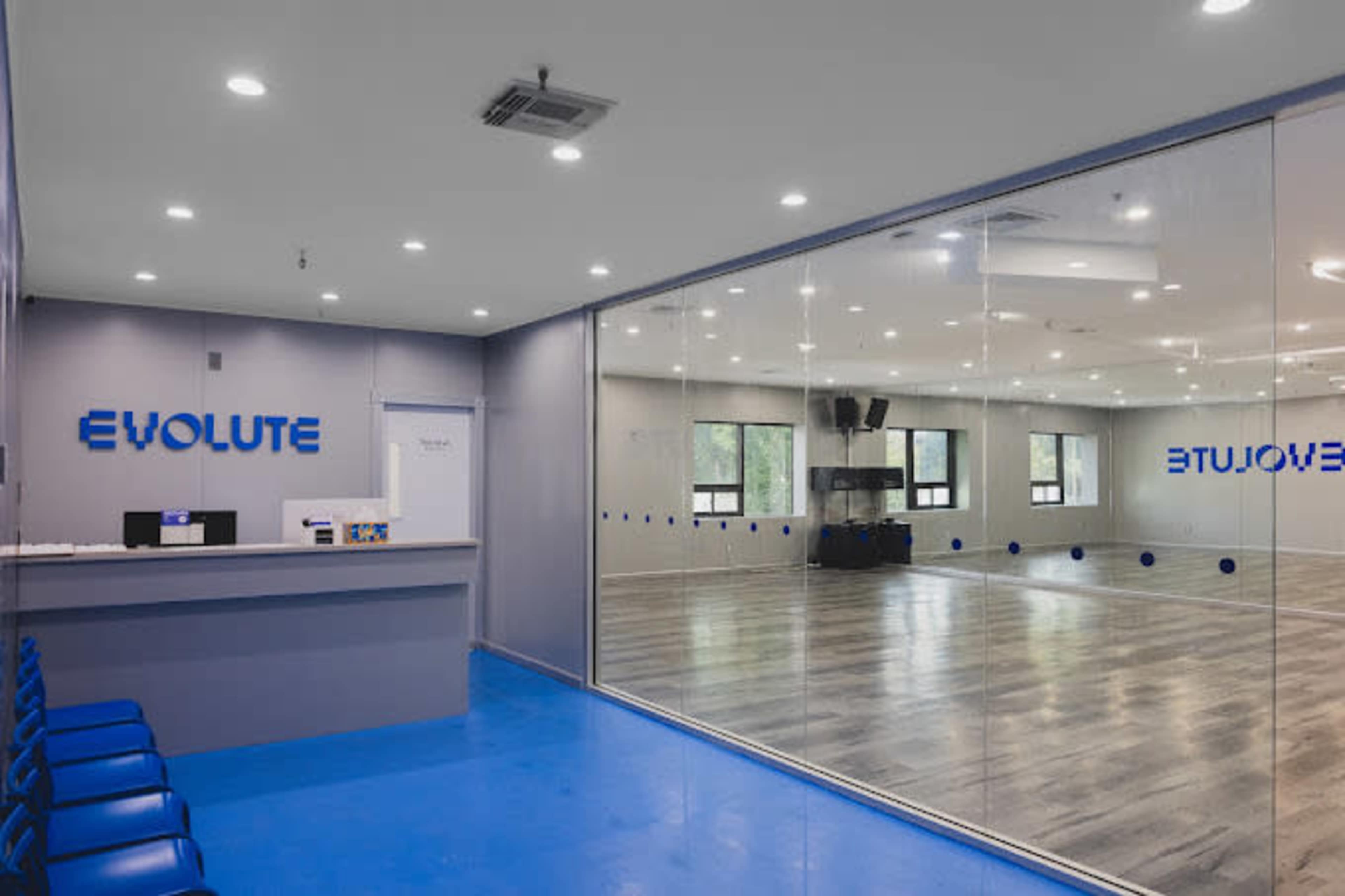 The image shows a dance studio with a reception area featuring a blue floor, a counter with the name "Evolute," and a large mirror on the wall reflecting the dance space.