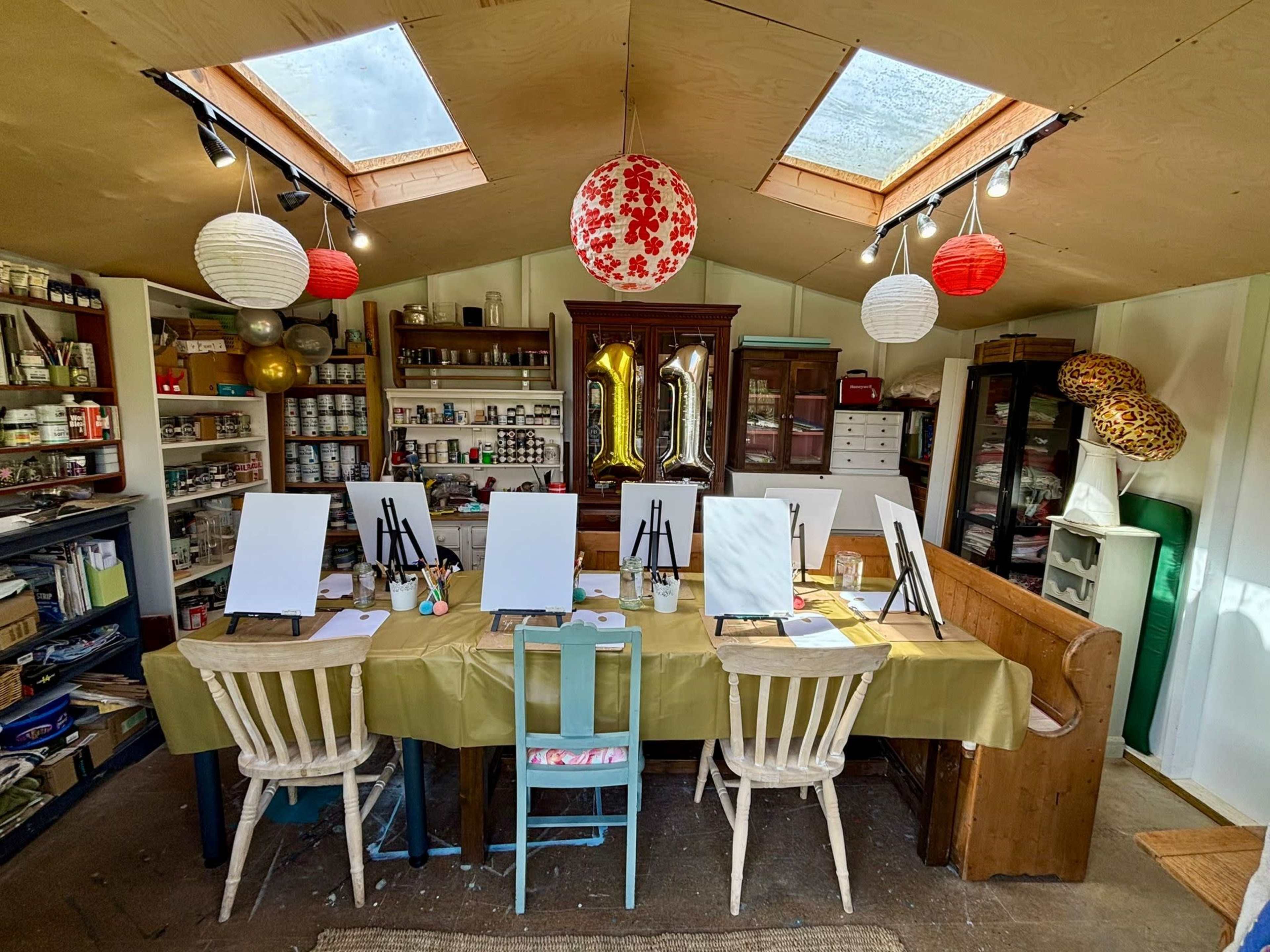 The image shows an art studio with a long table set up with blank canvases, chairs, and various art supplies, illuminated by natural light from skylights.