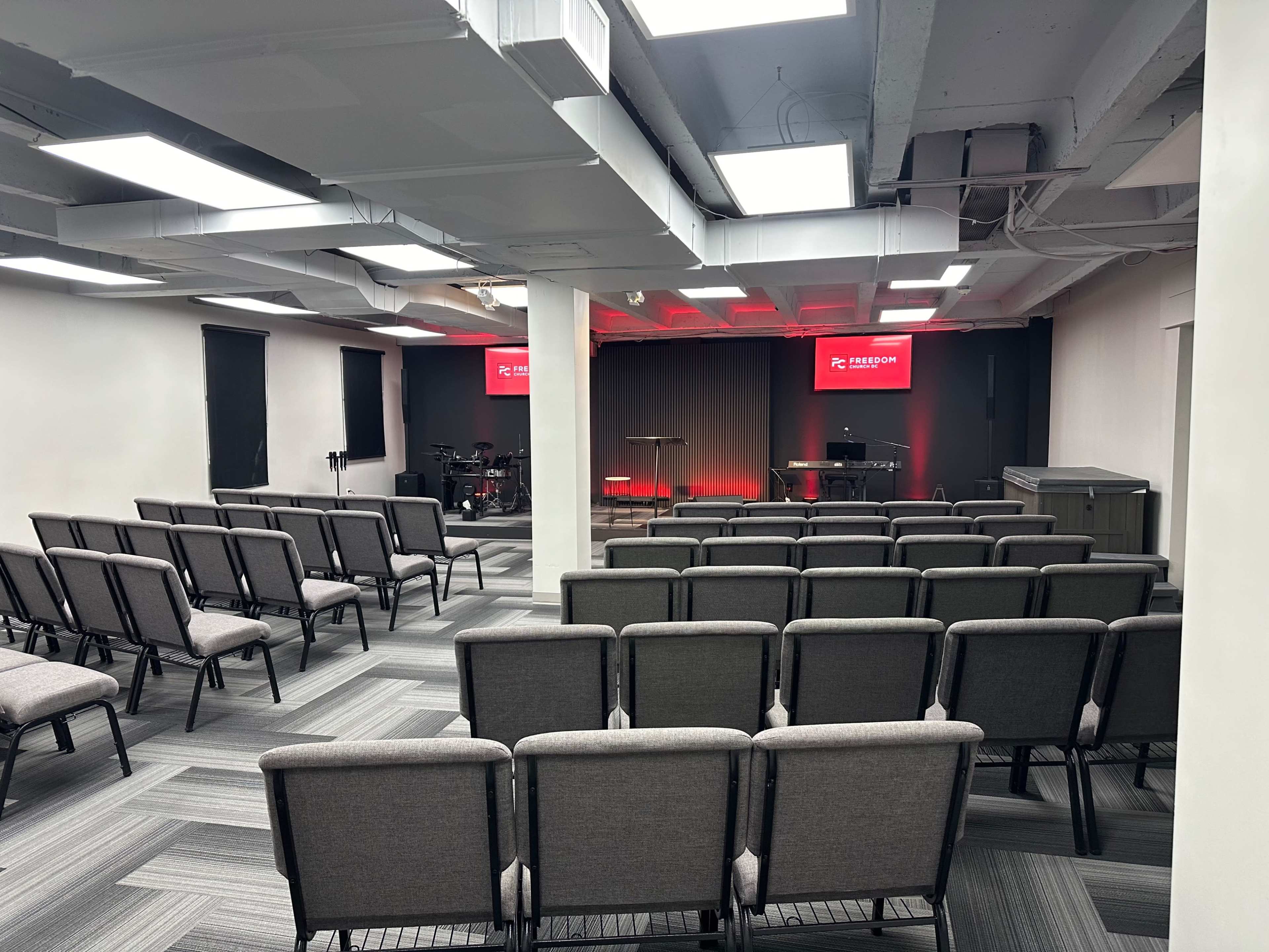 The image shows a spacious meeting room arranged with rows of gray chairs facing a small stage illuminated in red, with equipment and a screen visible in the background.