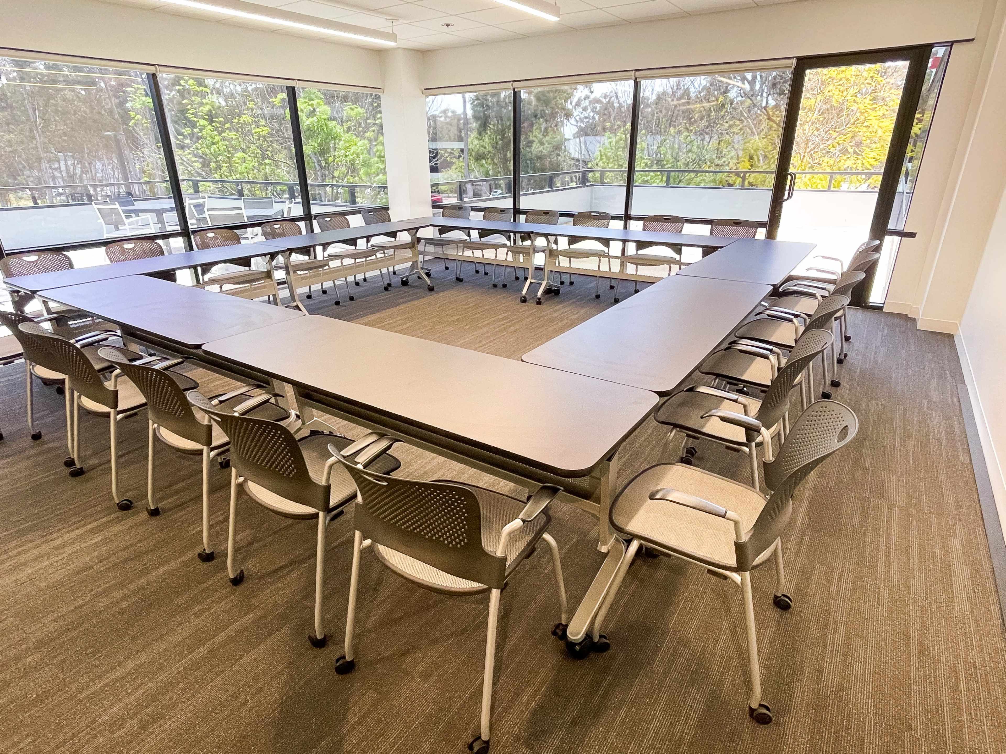 A conference room features a large, arranged table in a triangular shape with numerous chairs positioned around it, illuminated by natural light from large windows.