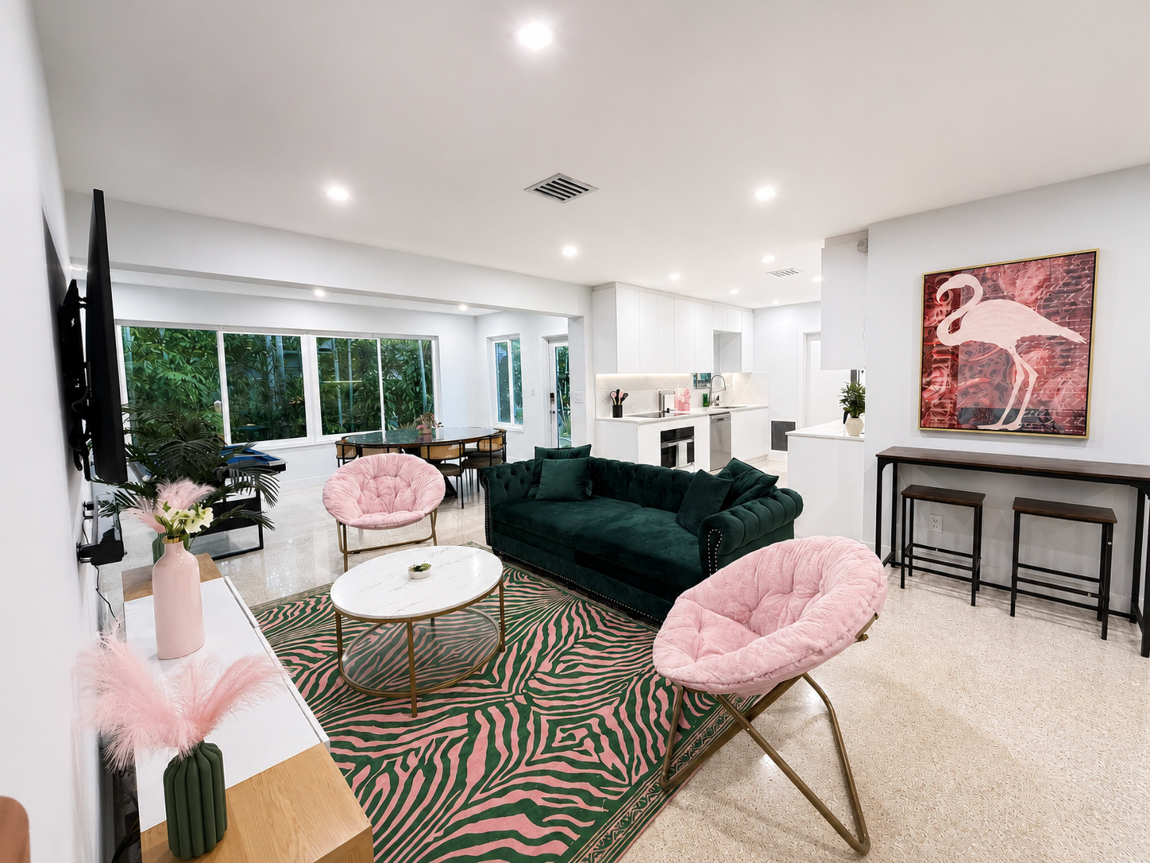 The living room features a green sofa, pink accent chairs, a round coffee table, and a colorful area rug, with large windows allowing natural light to enter.