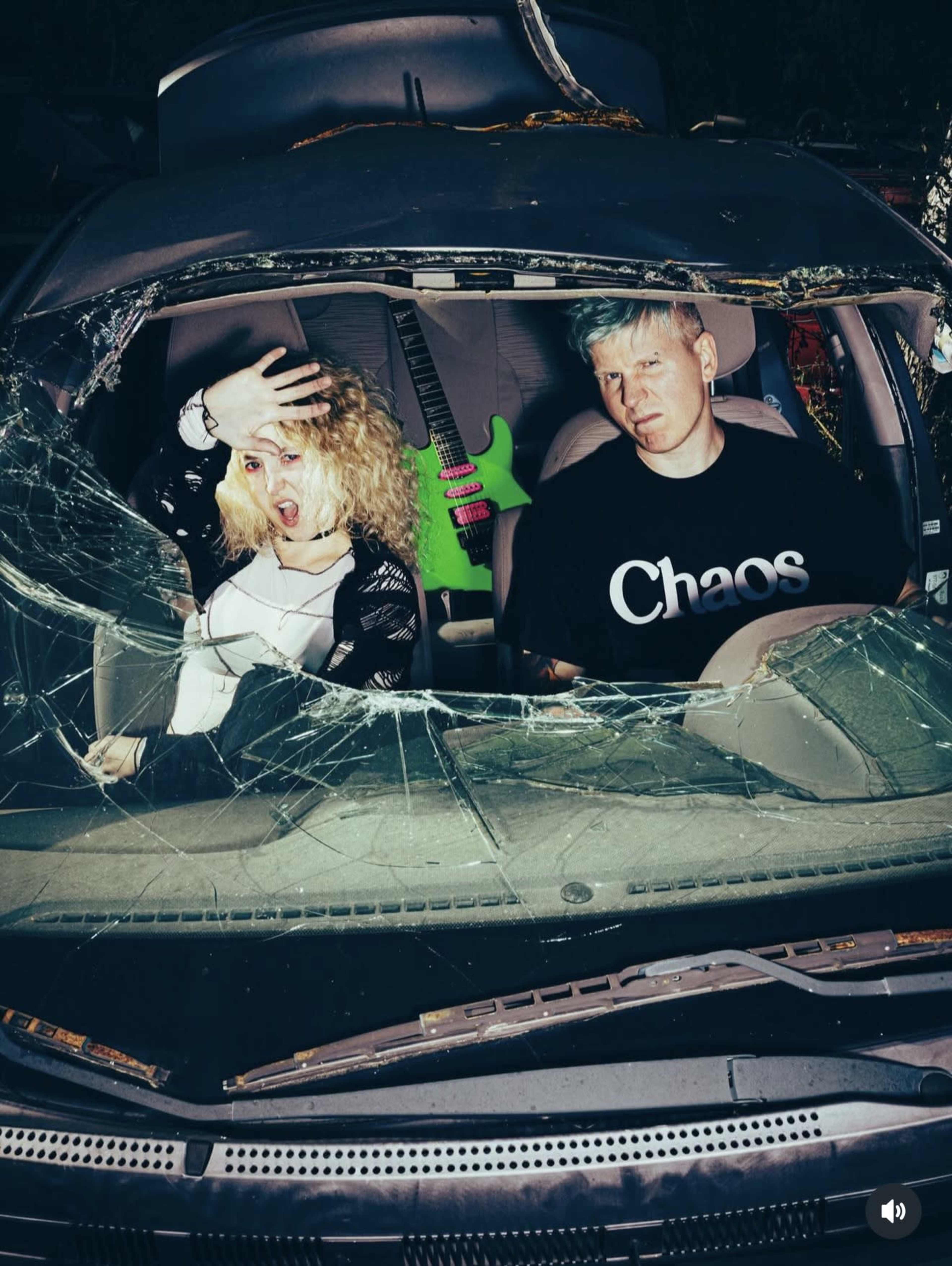 A woman with curly blonde hair and a man with blue hair sit inside a car with a shattered windshield, and a green guitar is visible in the background.