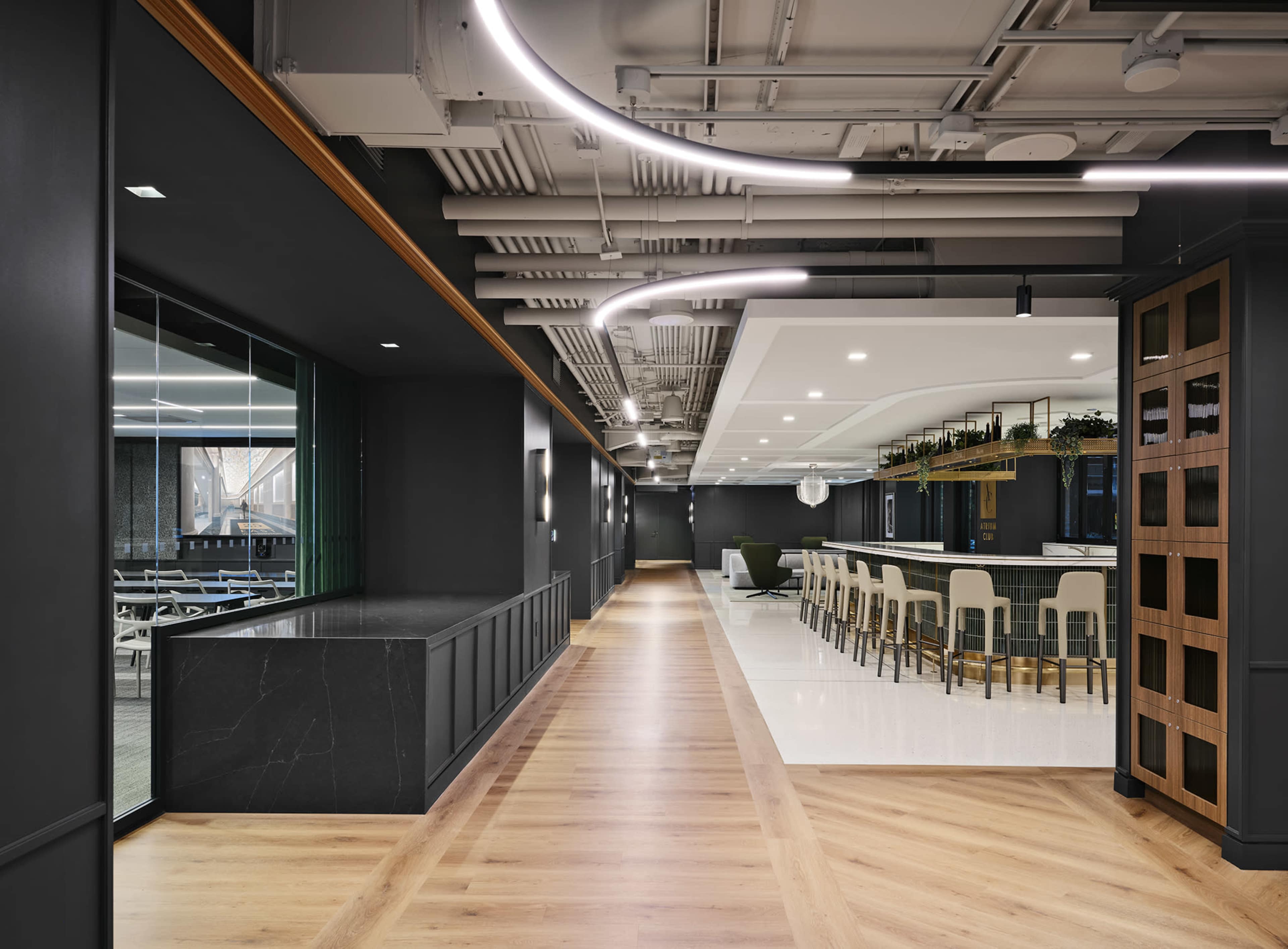 The image shows a modern office interior featuring a long hallway with a dark wall, a sleek bar area with high stools, and a spacious dining area with large windows.