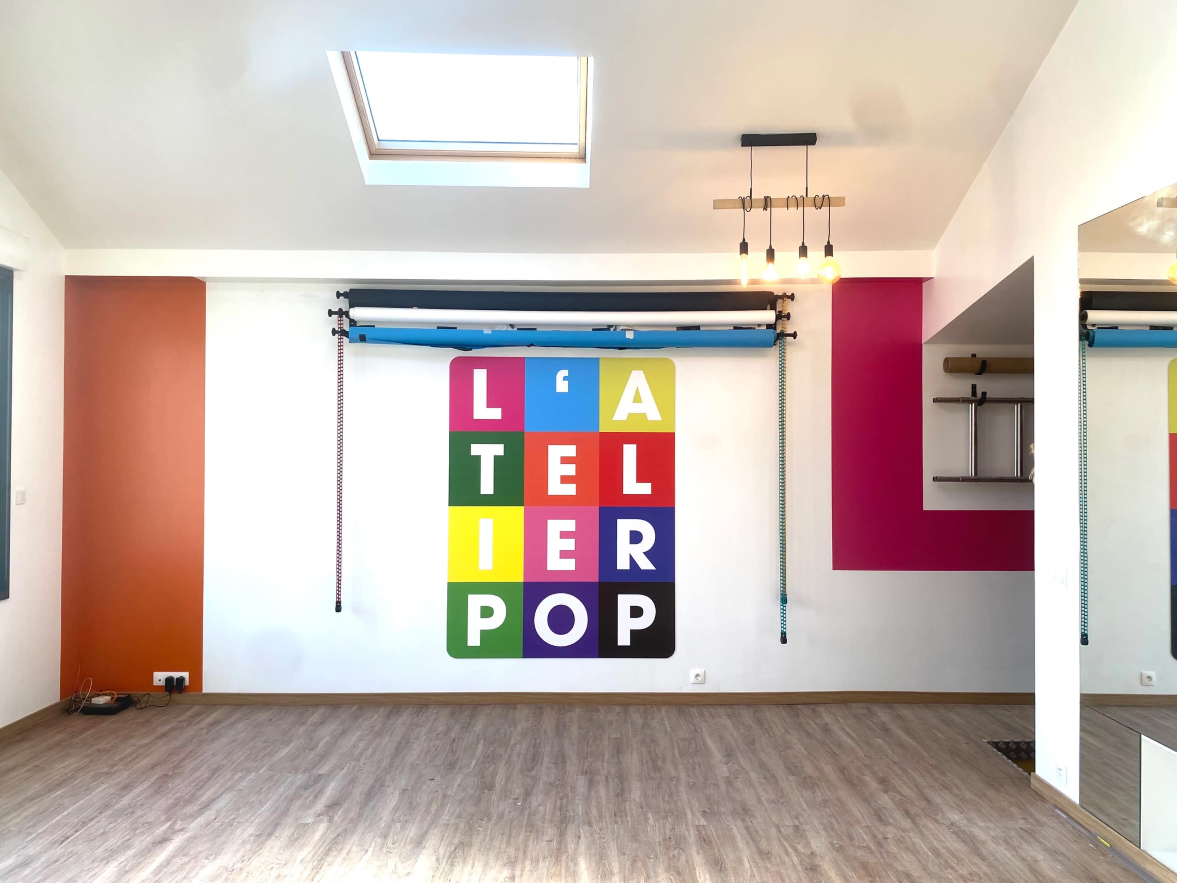 The image shows a bright, modern room featuring a colorful wall mural that spells out "L'ATELIER POP" with a skylight above and a ceiling-mounted track with equipment.