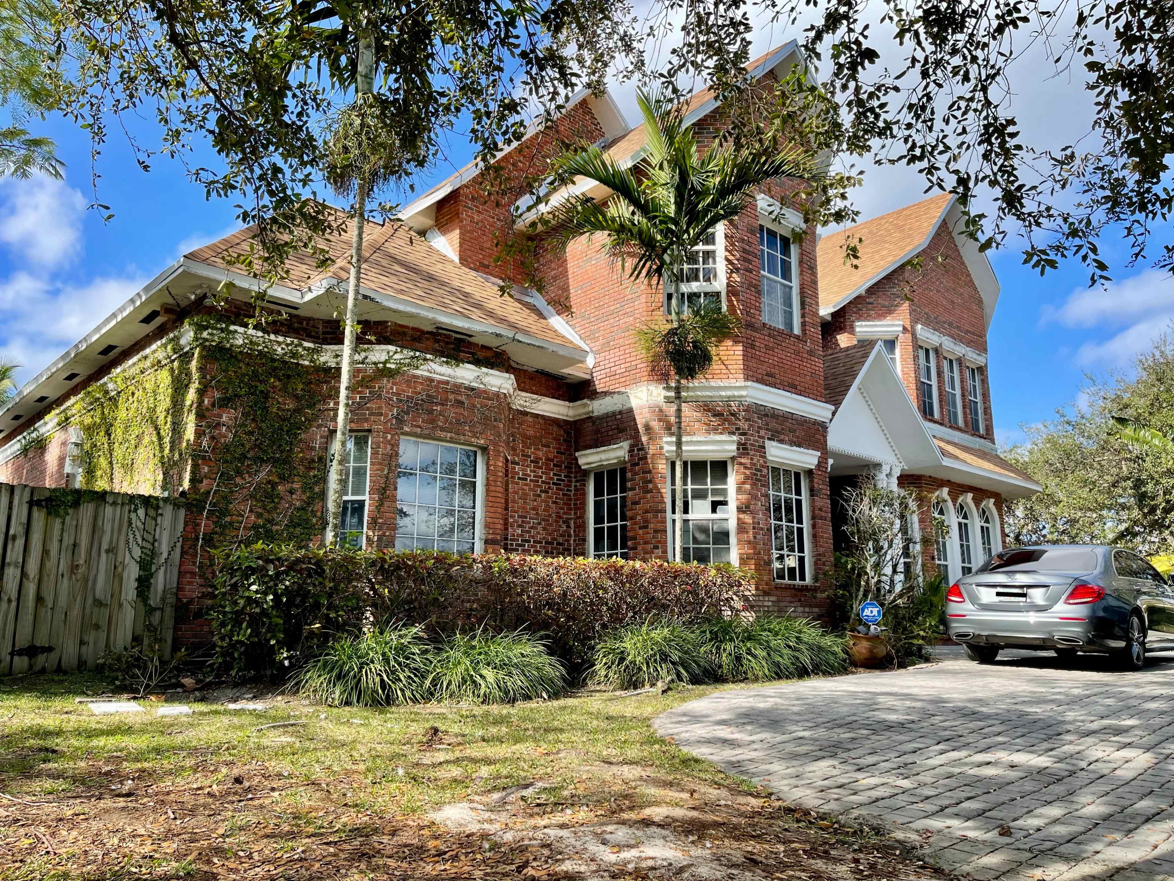 A large brick house with multiple gables and a paved driveway is surrounded by greenery and trees.