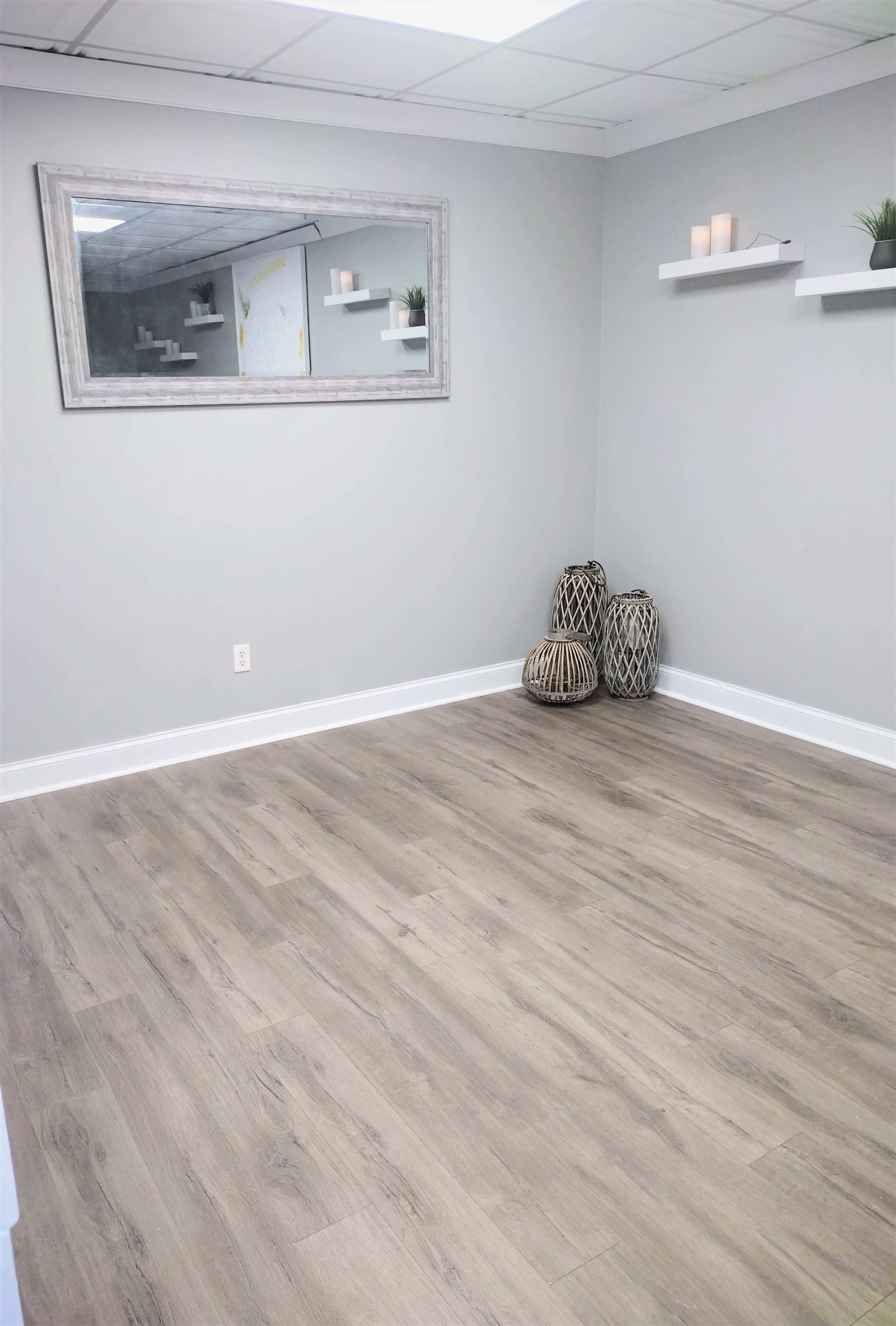 A sparsely furnished room with light-colored walls, a wooden floor, a large mirror on the wall, and decorative items arranged on shelves.