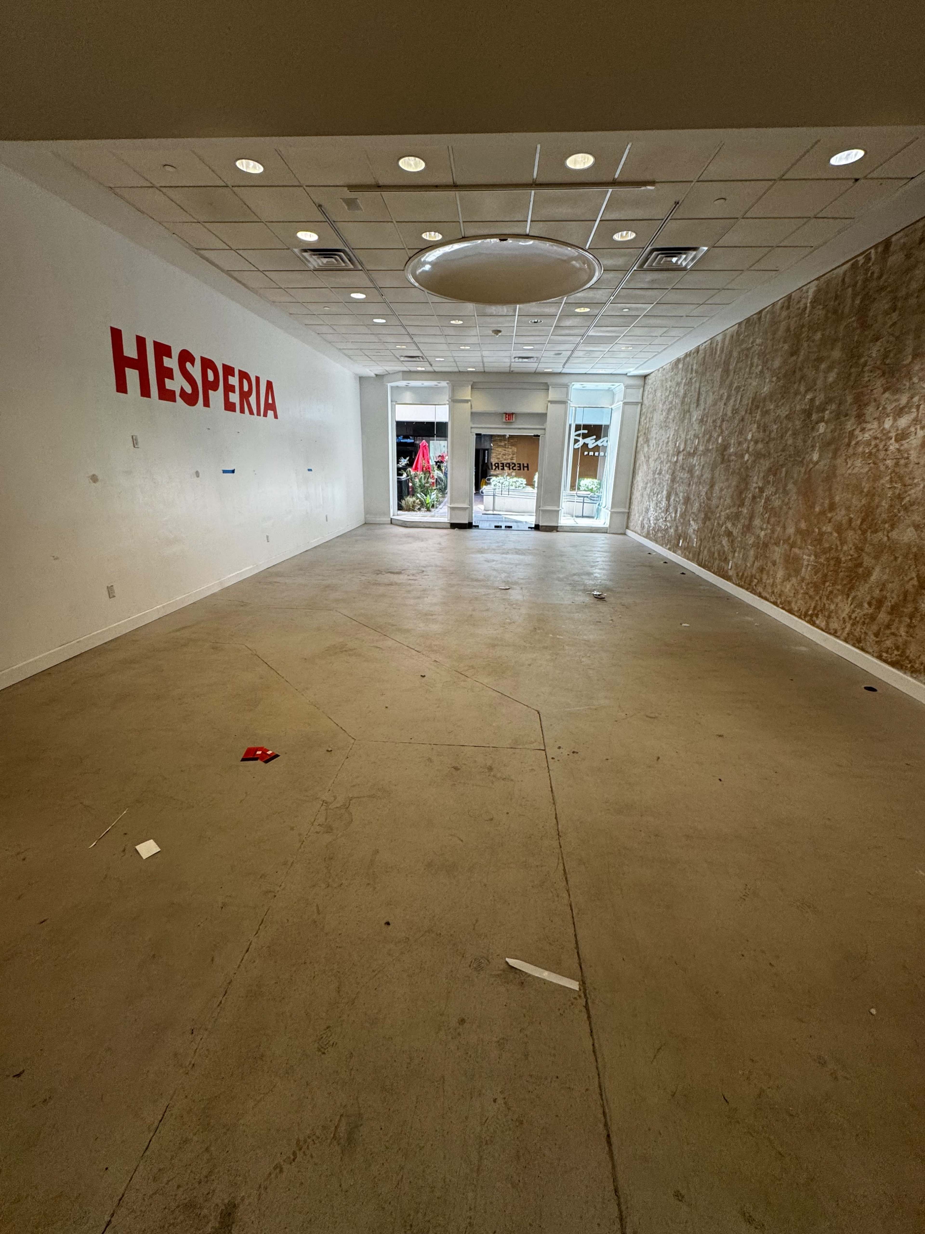The image shows a vacant interior space with white walls, a partially textured wall on one side, and the word "HESPERIA" displayed prominently.