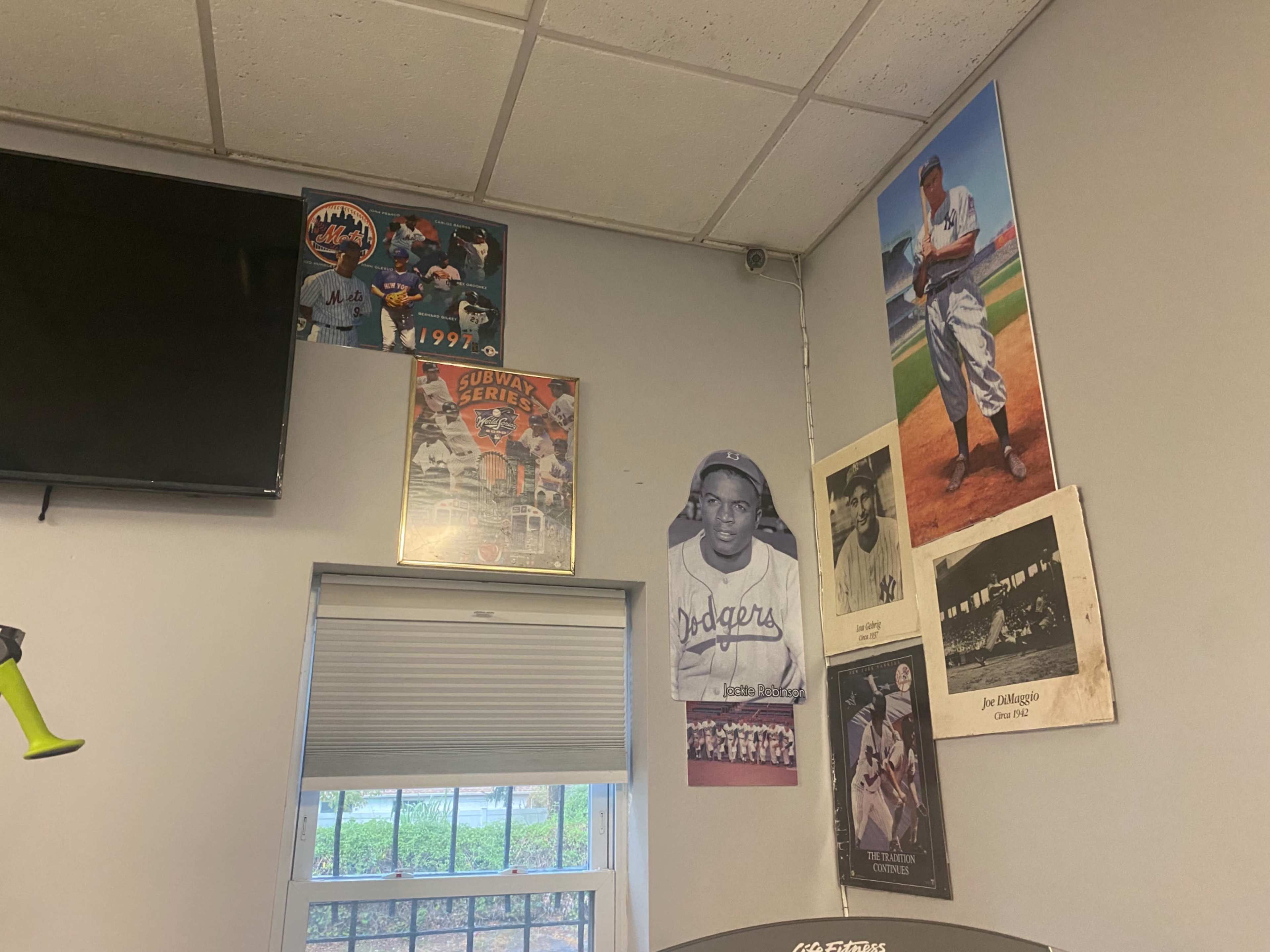 A corner of a room with a television, various sports posters on the walls, including one of a baseball player, and a window with a blind partially drawn.