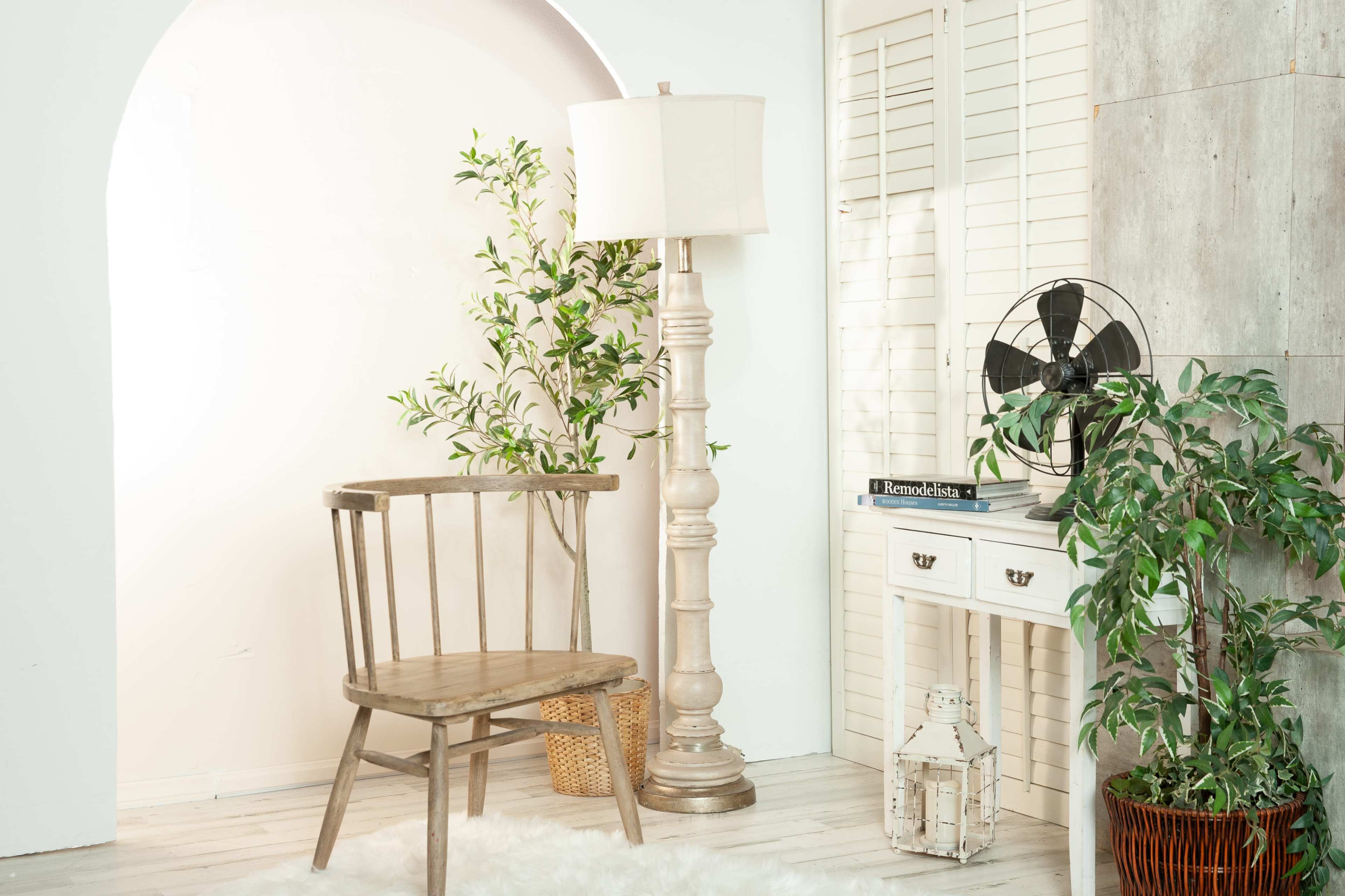 The scene features a wooden chair next to a decorative lamp, a potted plant, a vintage fan, and a white console table against a light-colored wall.