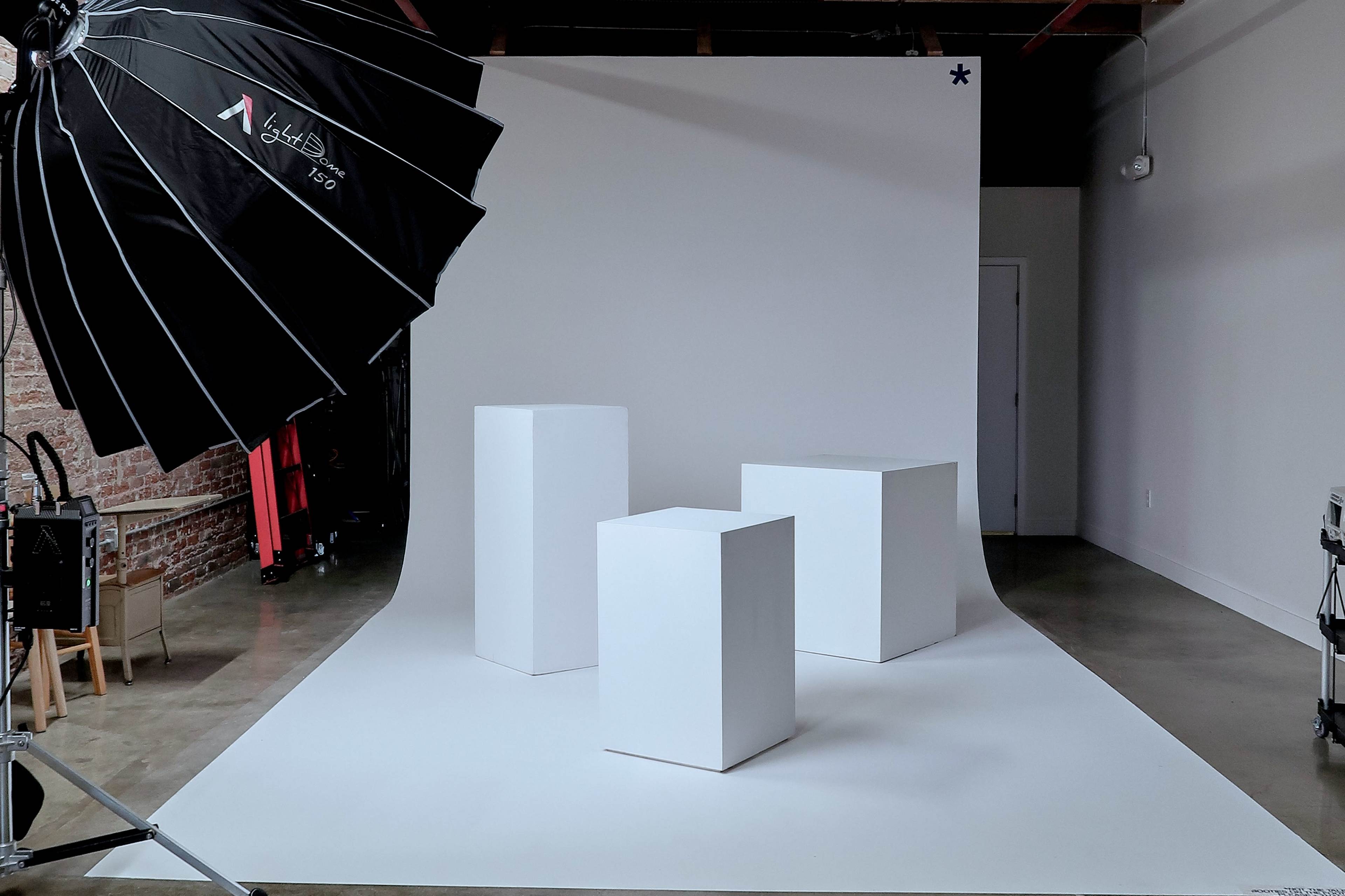 A photography studio features three white display pedestals on a seamless backdrop with a large softbox light positioned to the side.