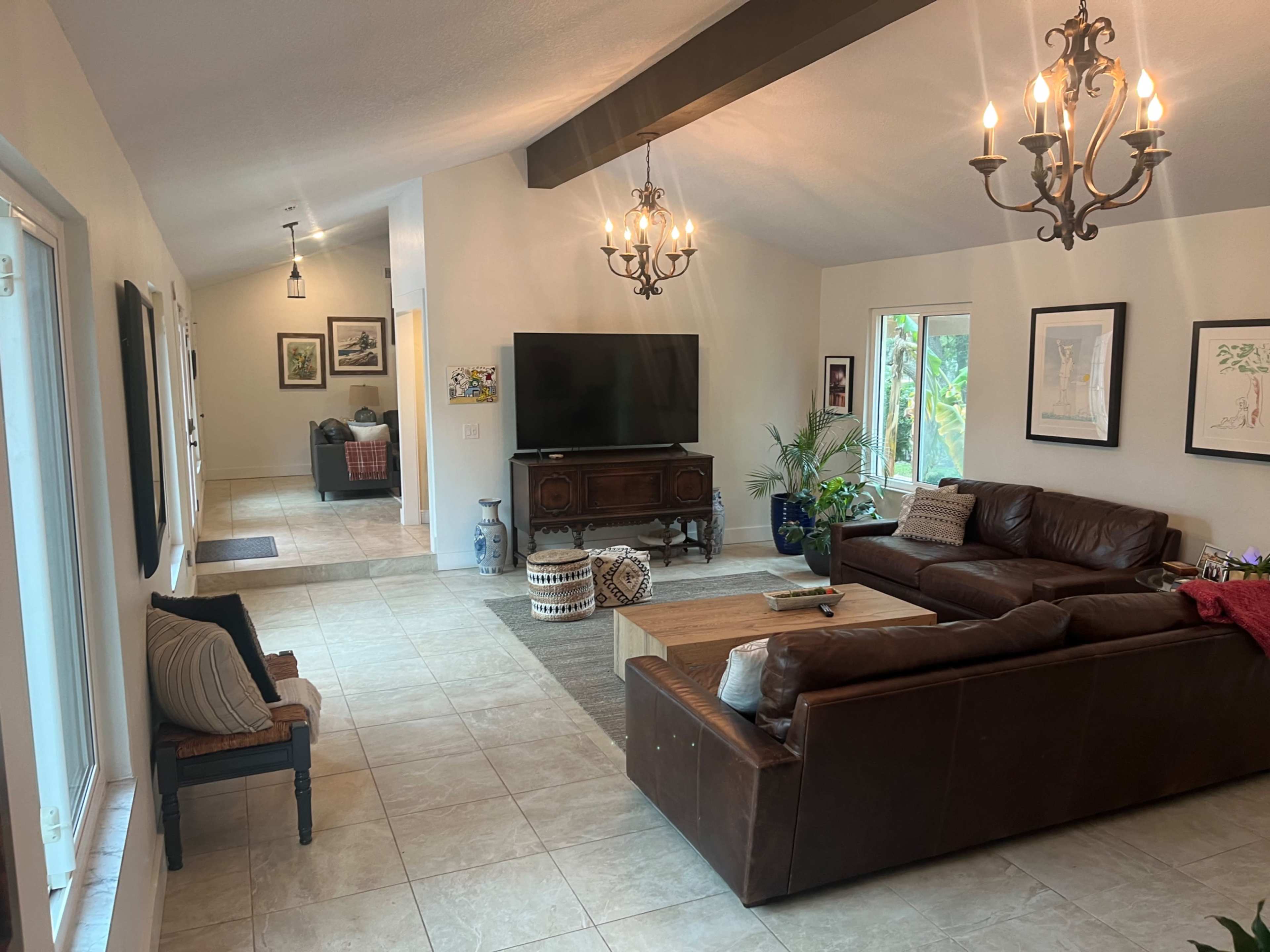 A spacious living room with a large sectional sofa, a wooden coffee table, a television, and large windows allowing natural light, along with a dining area visible in the background.