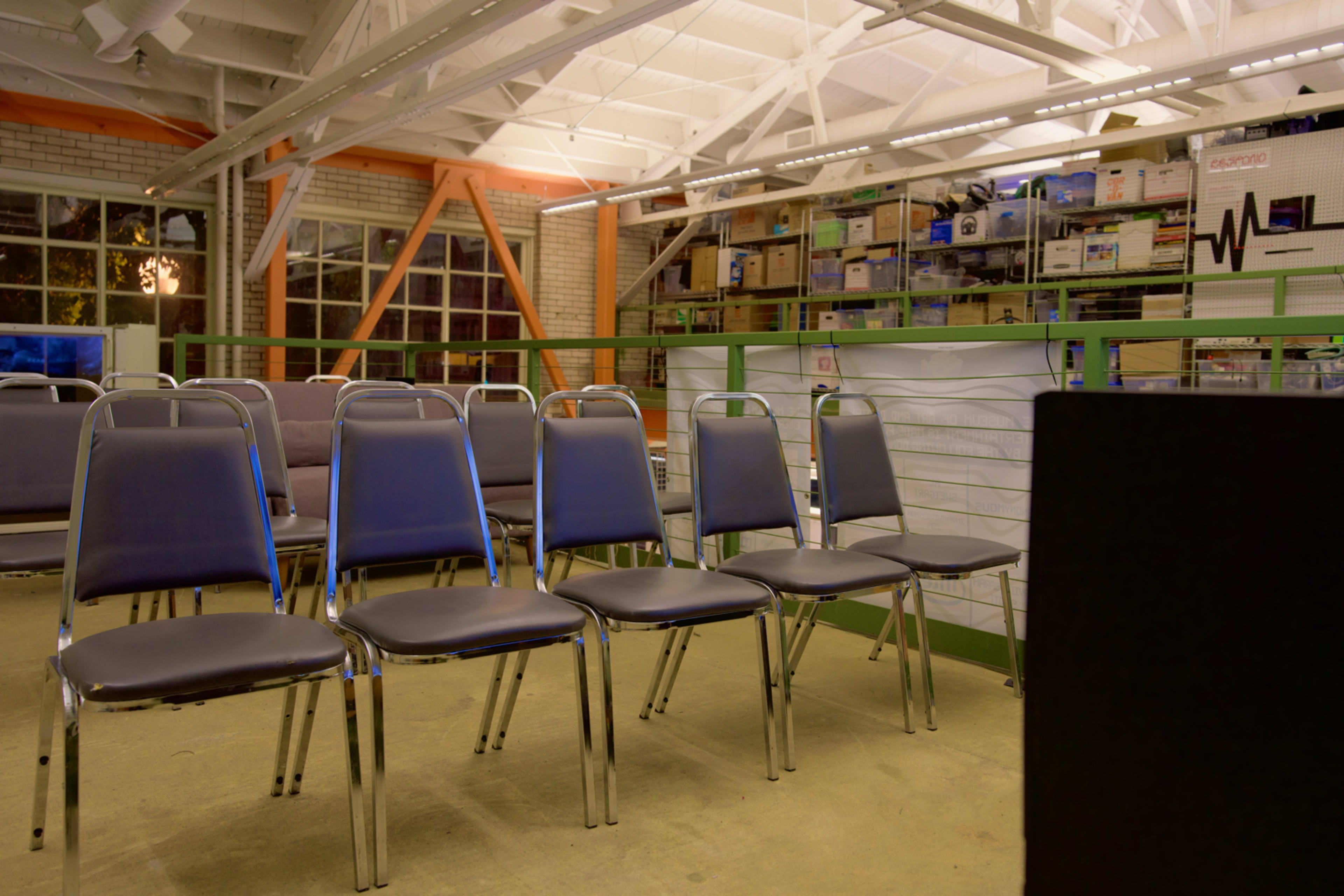A row of metal chairs is lined up in a brightly lit, industrial-style room filled with shelves of boxes.