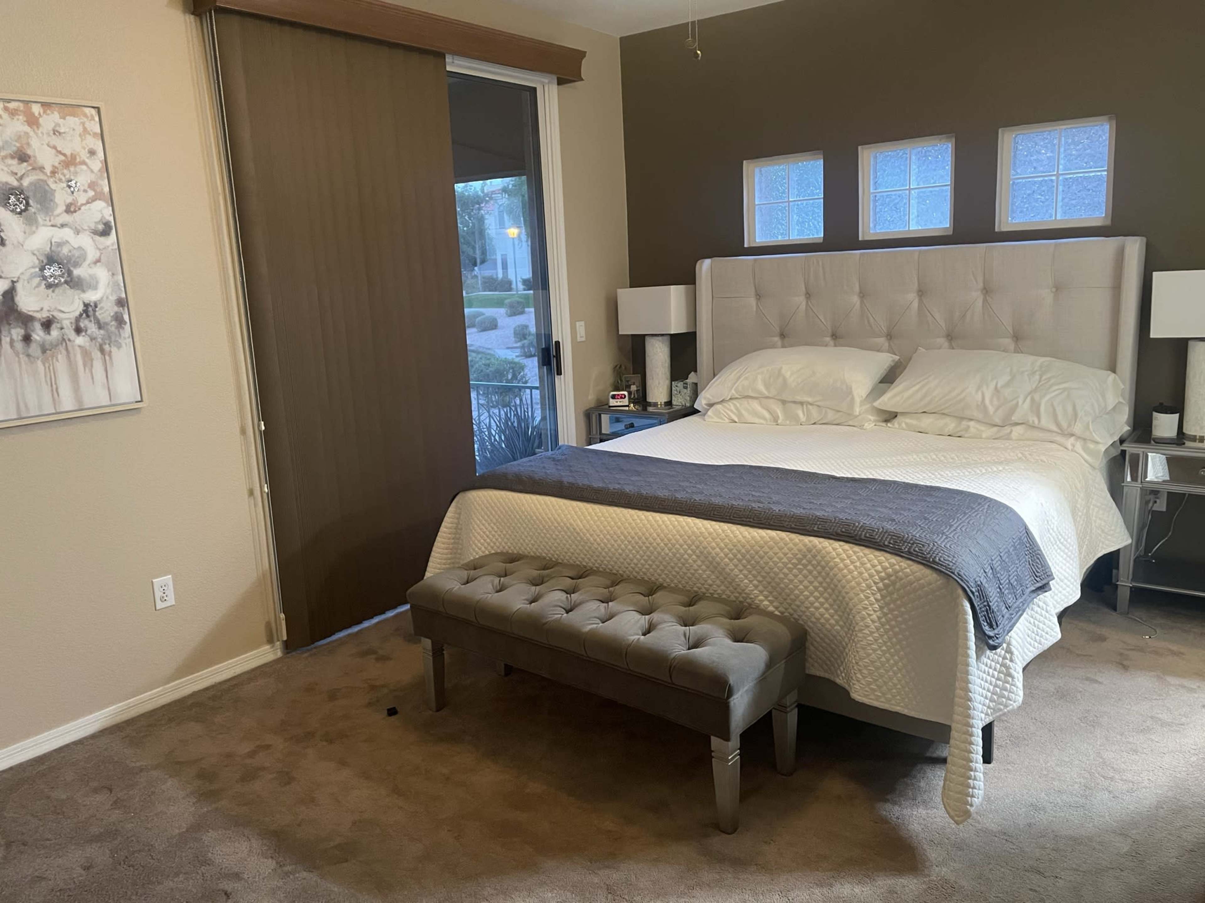 The image shows a bedroom with a bed, a bench at the foot, and sliding glass doors leading to an outdoor area.