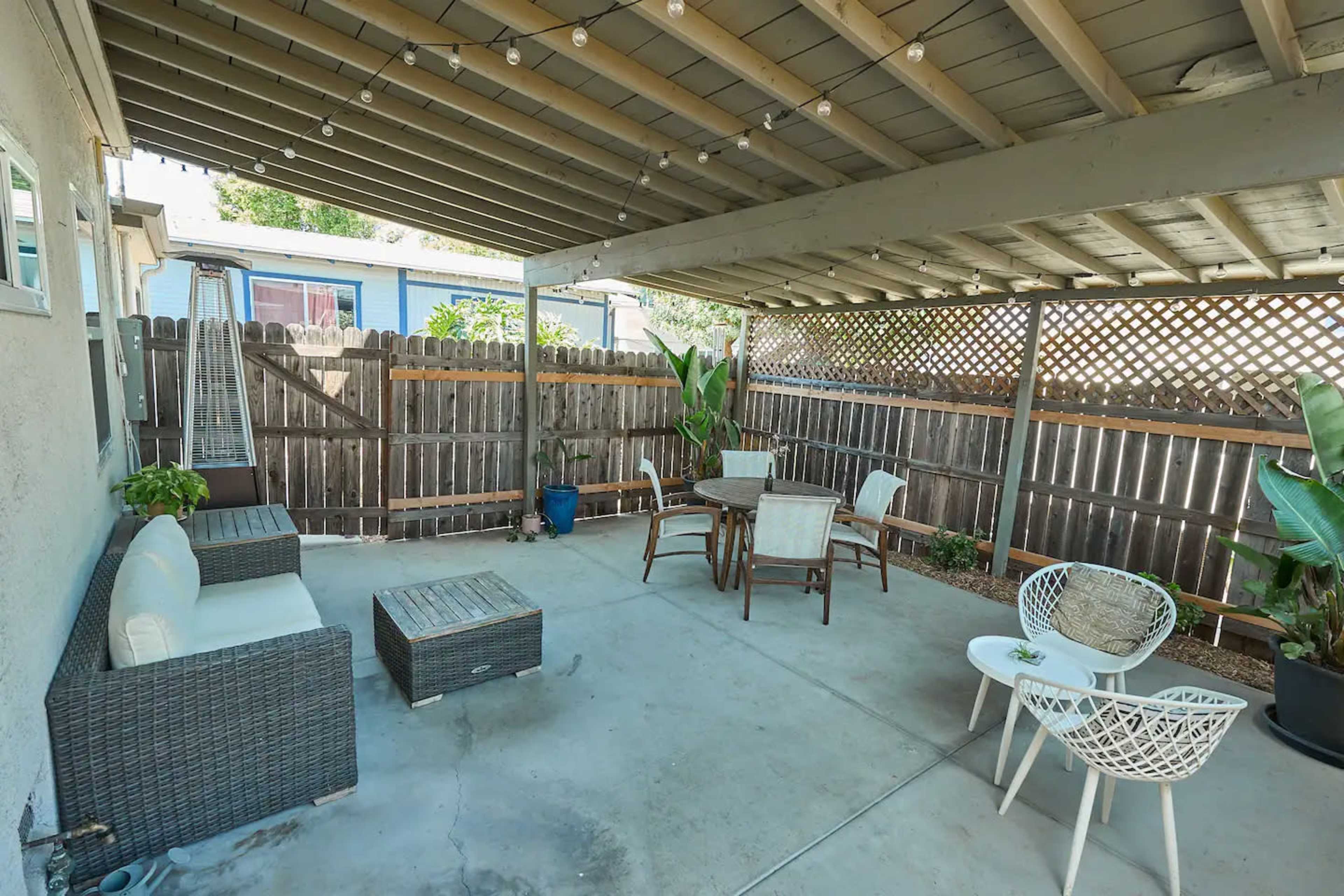 The image shows a covered outdoor seating area with a mix of wicker furniture, a small table, and a wooden fence in the background.