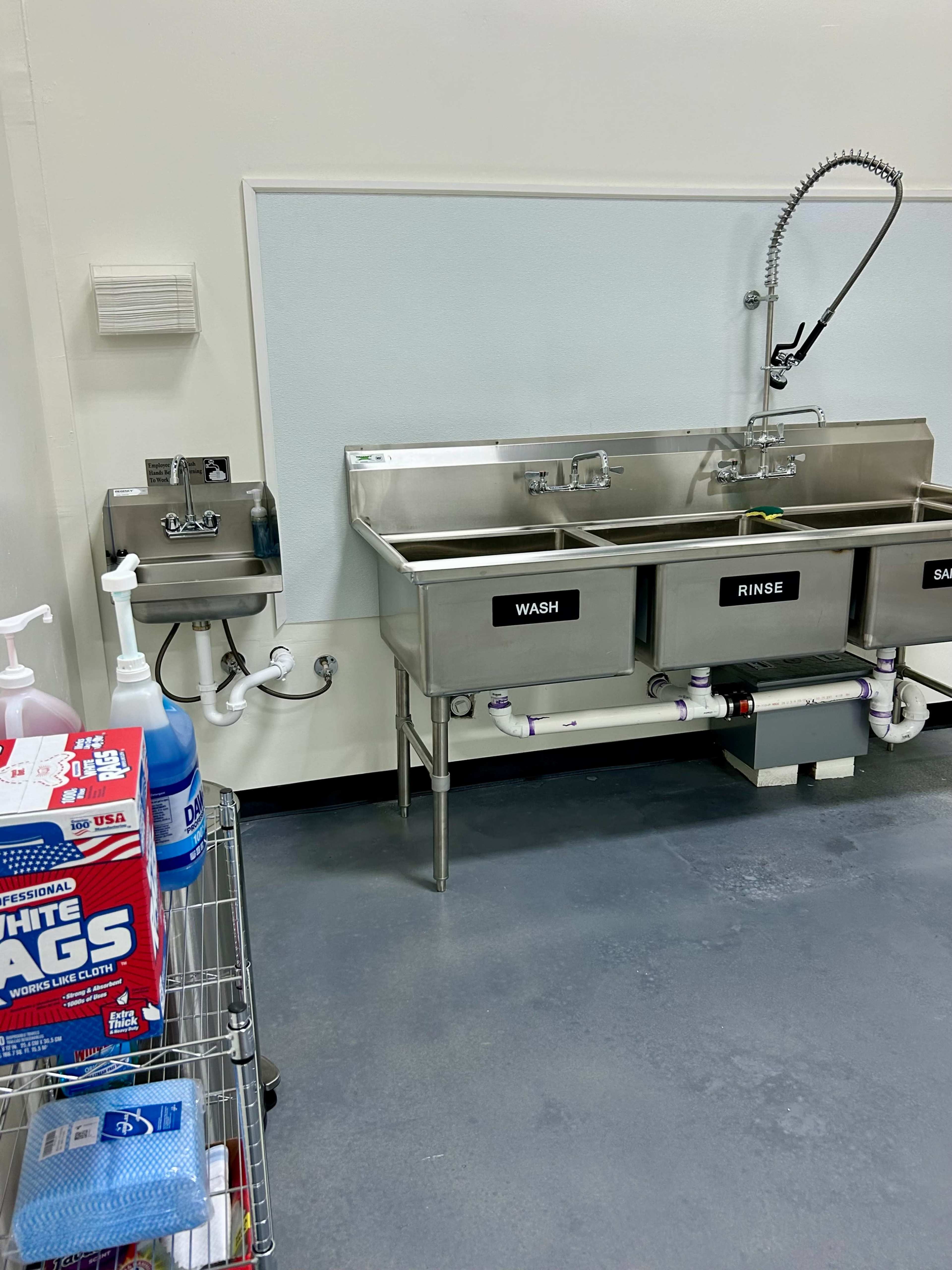The image shows a stainless steel three-compartment sink labeled "WASH," "RINSE," and "SANITIZE," alongside a cart with cleaning supplies and a sanitizer dispenser on the wall.